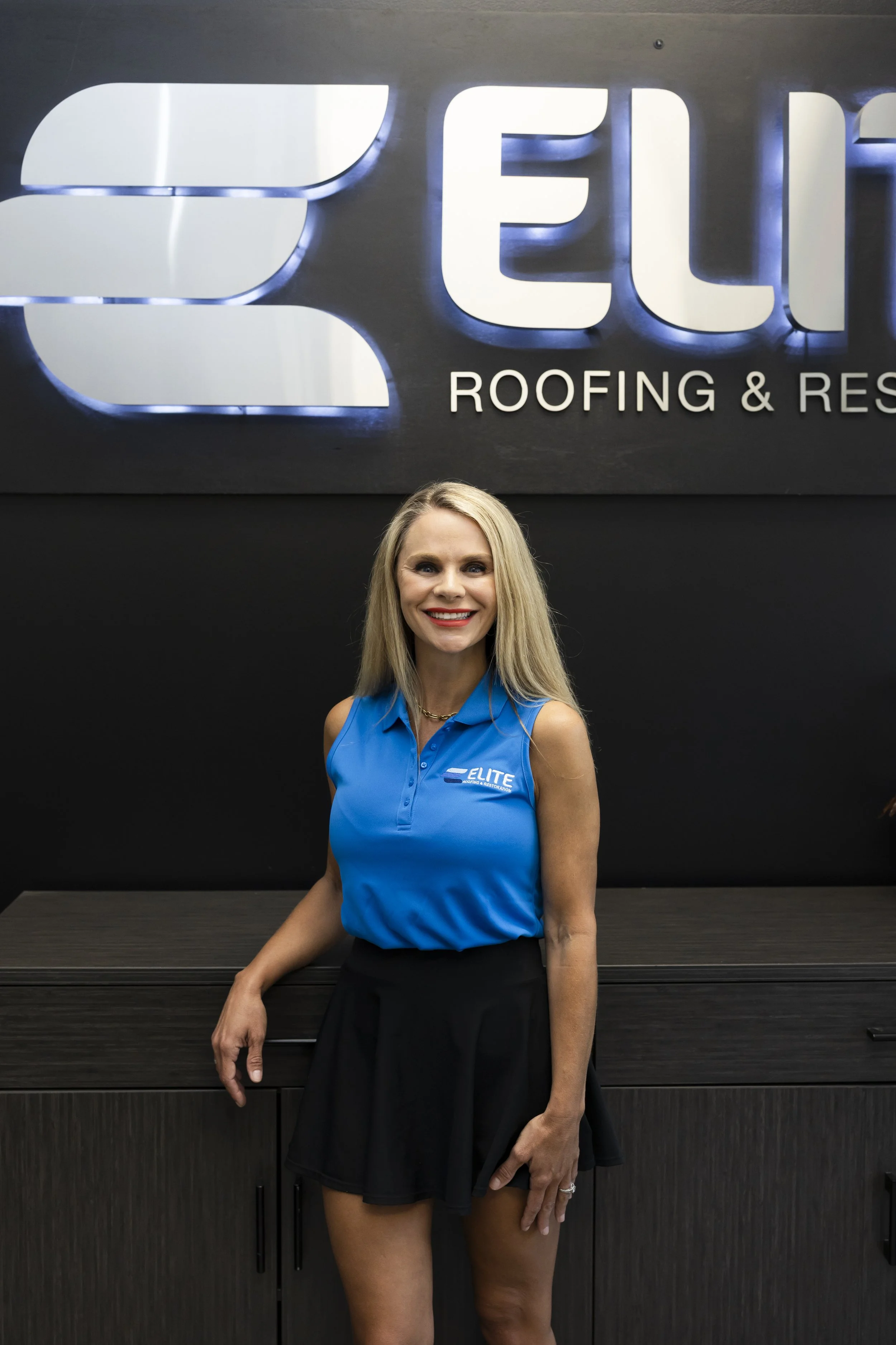 A smiling woman with long blonde hair wearing a blue polo shirt, standing in front of a wall decorated with small American flags.