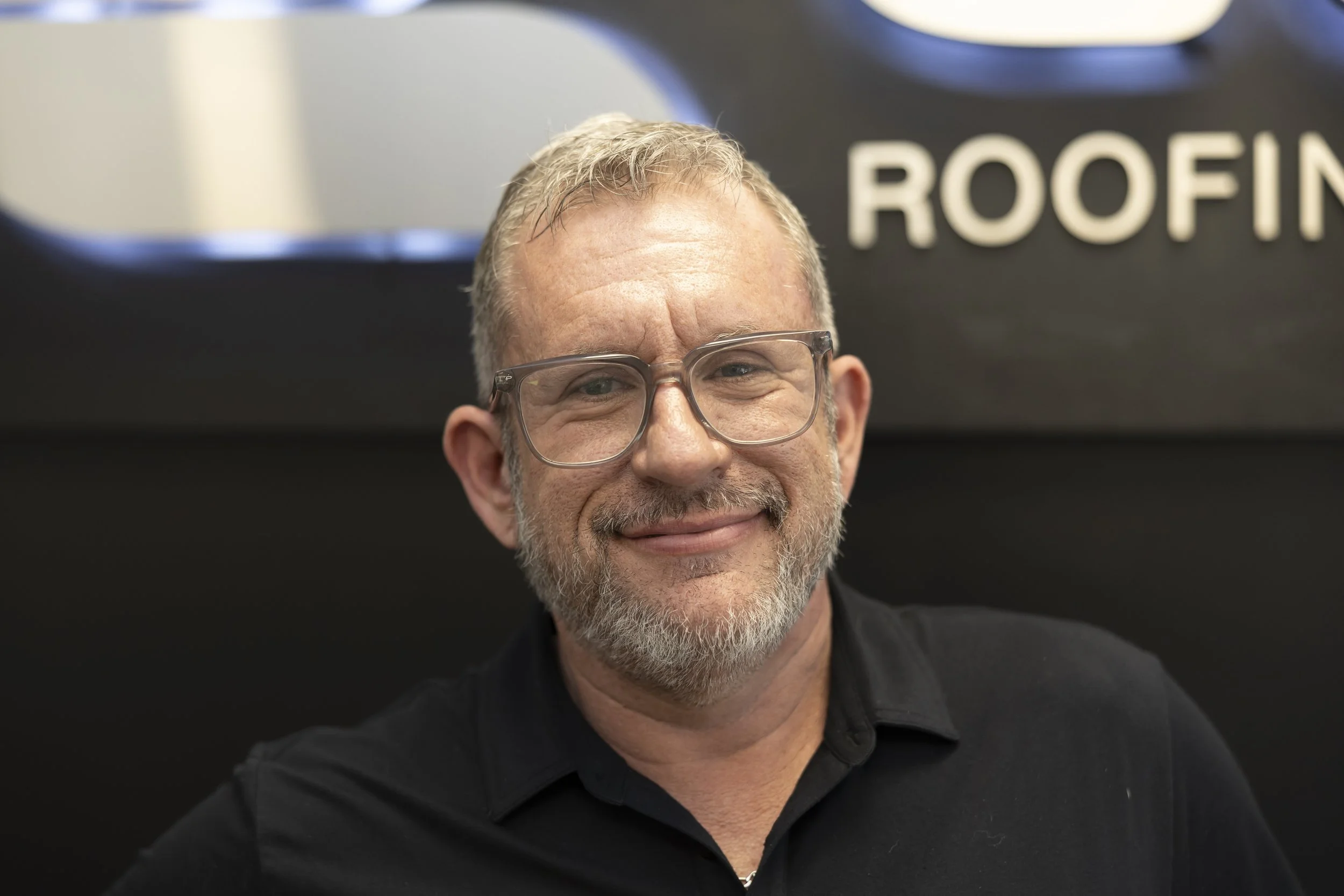 A smiling man with gray hair and beard wearing a gray polo shirt with an 'ELITE' logo, sitting indoors near a window with a meal and drink on the table behind him.