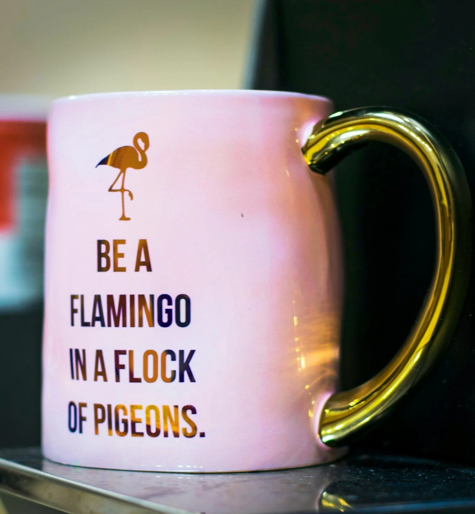Close-up of a white ceramic mug with a flamingo graphic and the words 'BE A FLAMINGO IN A FLOCK OF PIGEONS' printed on it. The mug has a gold-colored handle. The mug is placed on a silver dish.