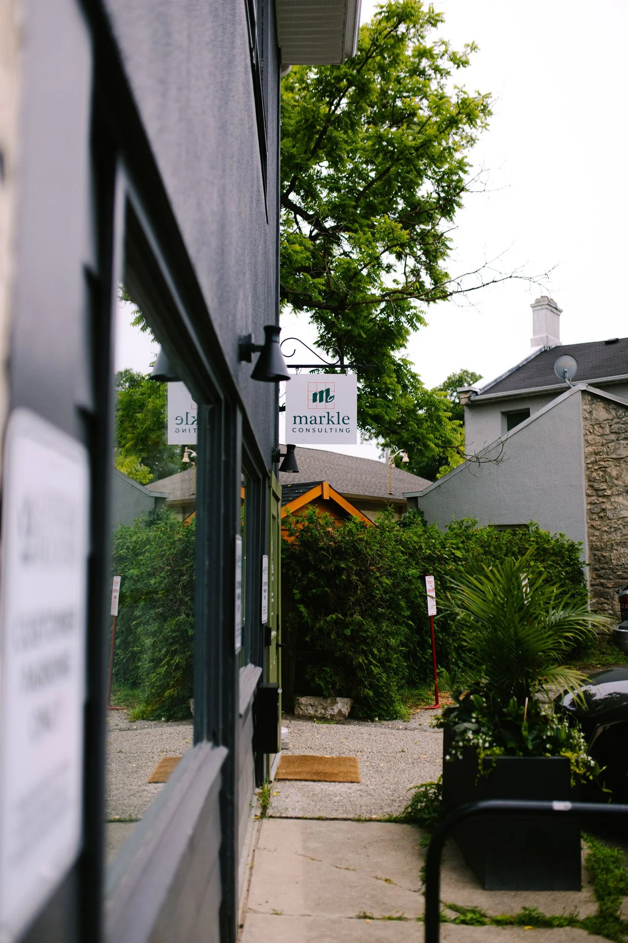View of a narrow sidewalk between a building and a lush garden, with a sign for 'Markle Consulting' hanging above. The scene includes greenery, trees, residential houses, and parked vehicles in the background.