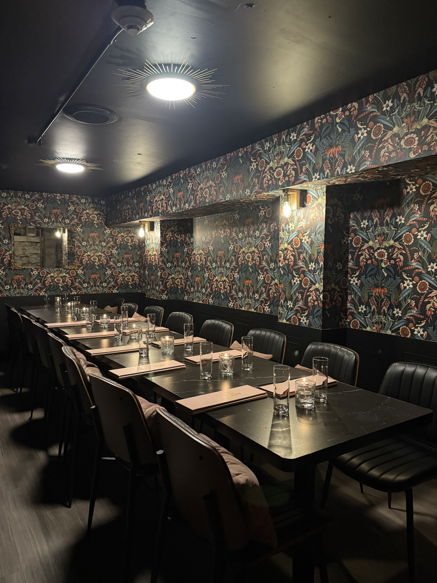 A dimly lit restaurant with a long black table set with glasses, napkins, and candles, surrounded by black chairs and decorated with floral wallpaper on the walls.