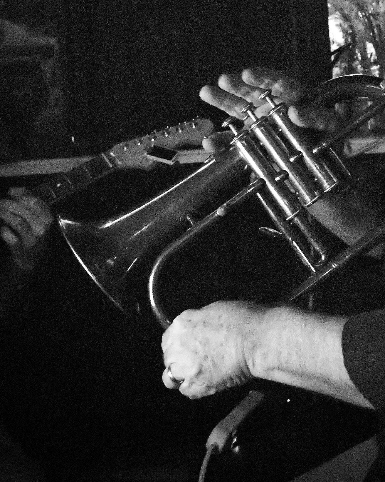 A close-up black and white photo of a person's hand holding a guitar and another hand pressing the strings on the fretboard, with a metallic instrument or object nearby.