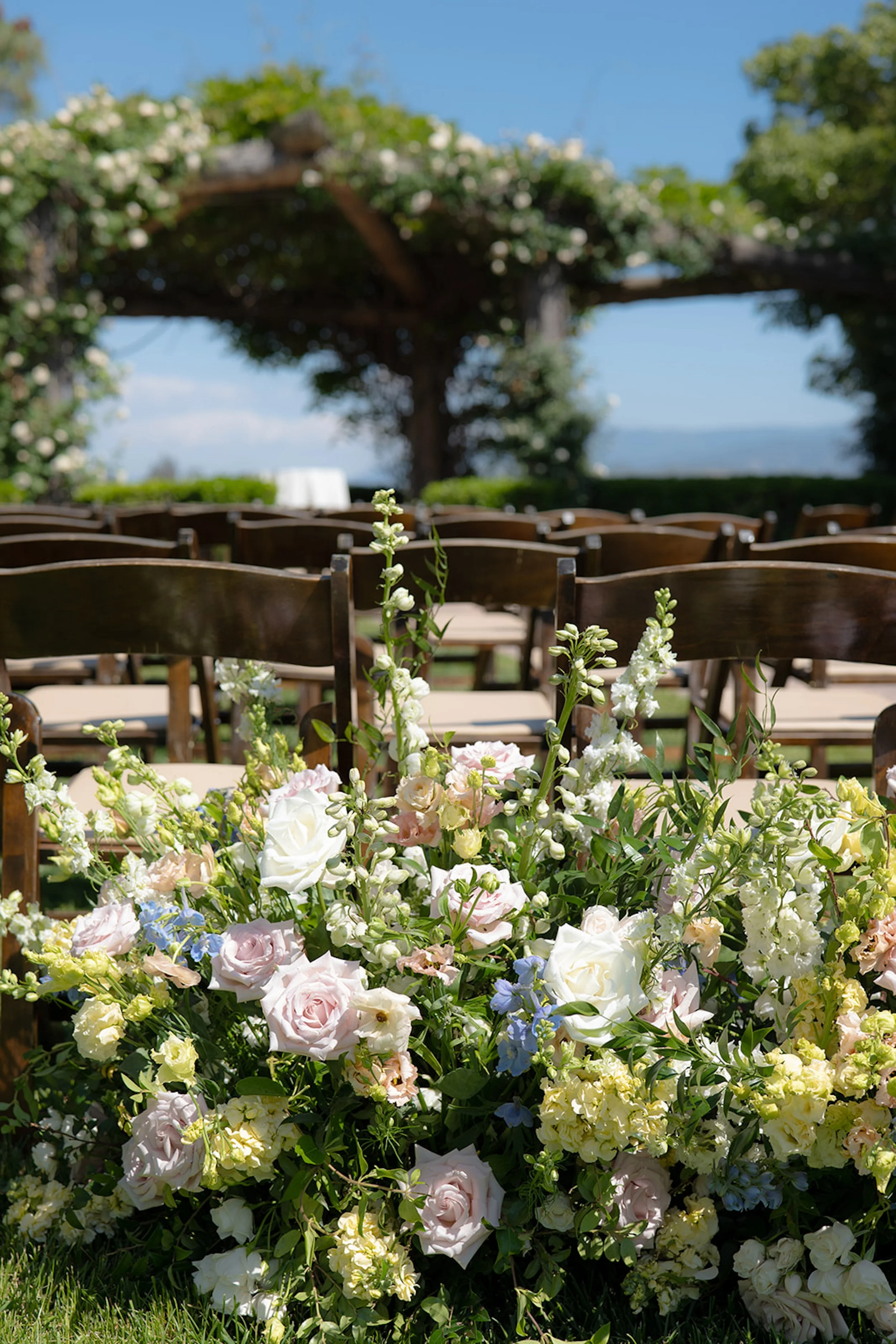Wedding ceremony setup with rows of wooden chairs and a large floral arrangement of white, pink, and blue flowers in the foreground, under a tree with white blossoms. A South Coast Winery Wedding.