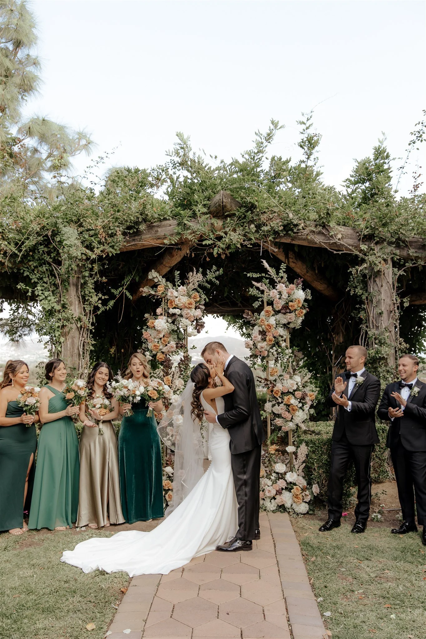 A wedding ceremony with a bride and groom kissing under an arch decorated with flowers, surrounded by bridesmaids and groomsmen outdoors. A South Coast Winery Wedding.