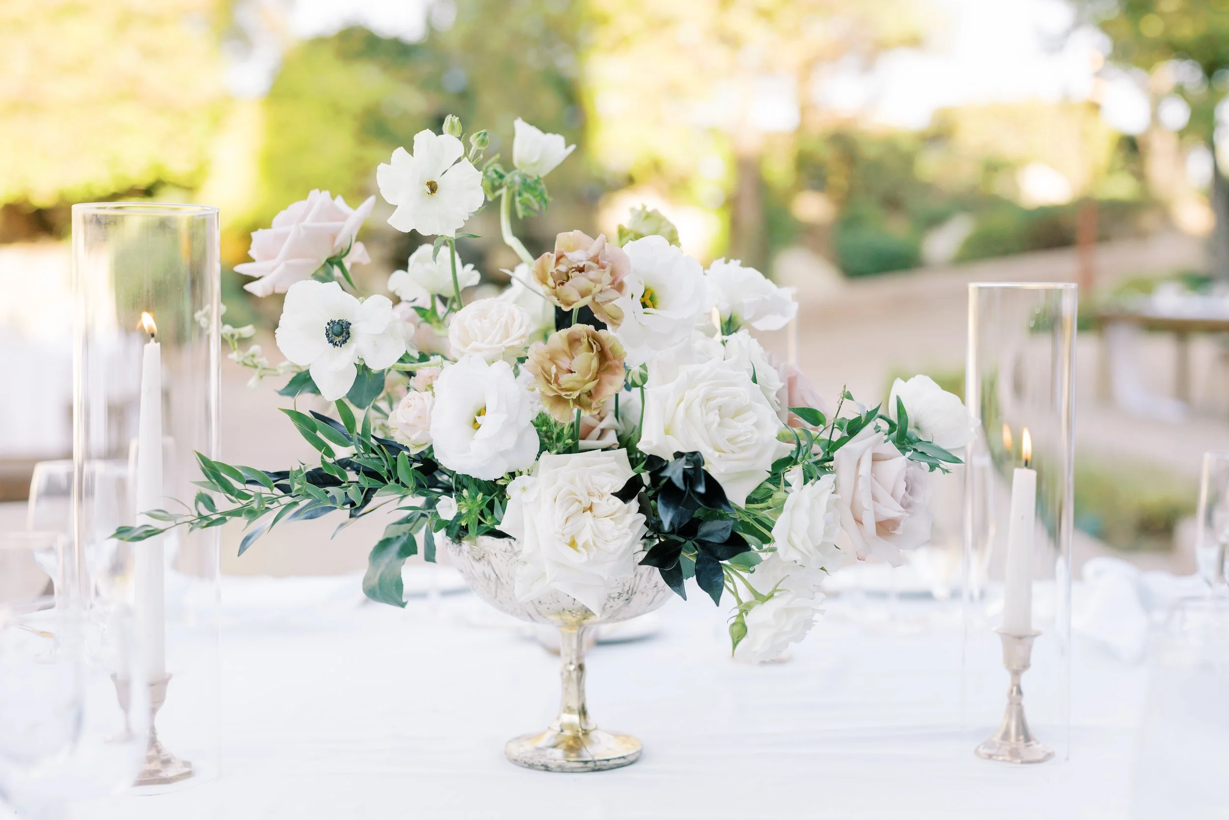tabletop image of floral centerpiece with white roses, toffee colored roses, anemones, in gold vase and gold tapered candle sticks with white candles and hurricane covers
