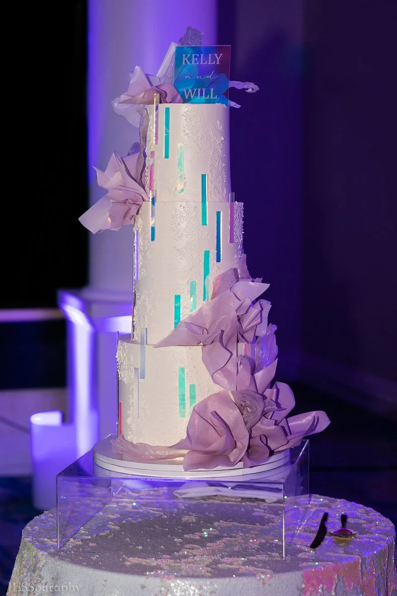 A tall white wedding cake decorated with light purple tissue paper flowers and illuminated by purple and blue lighting. A small cake topper reads "Kelly and Will."