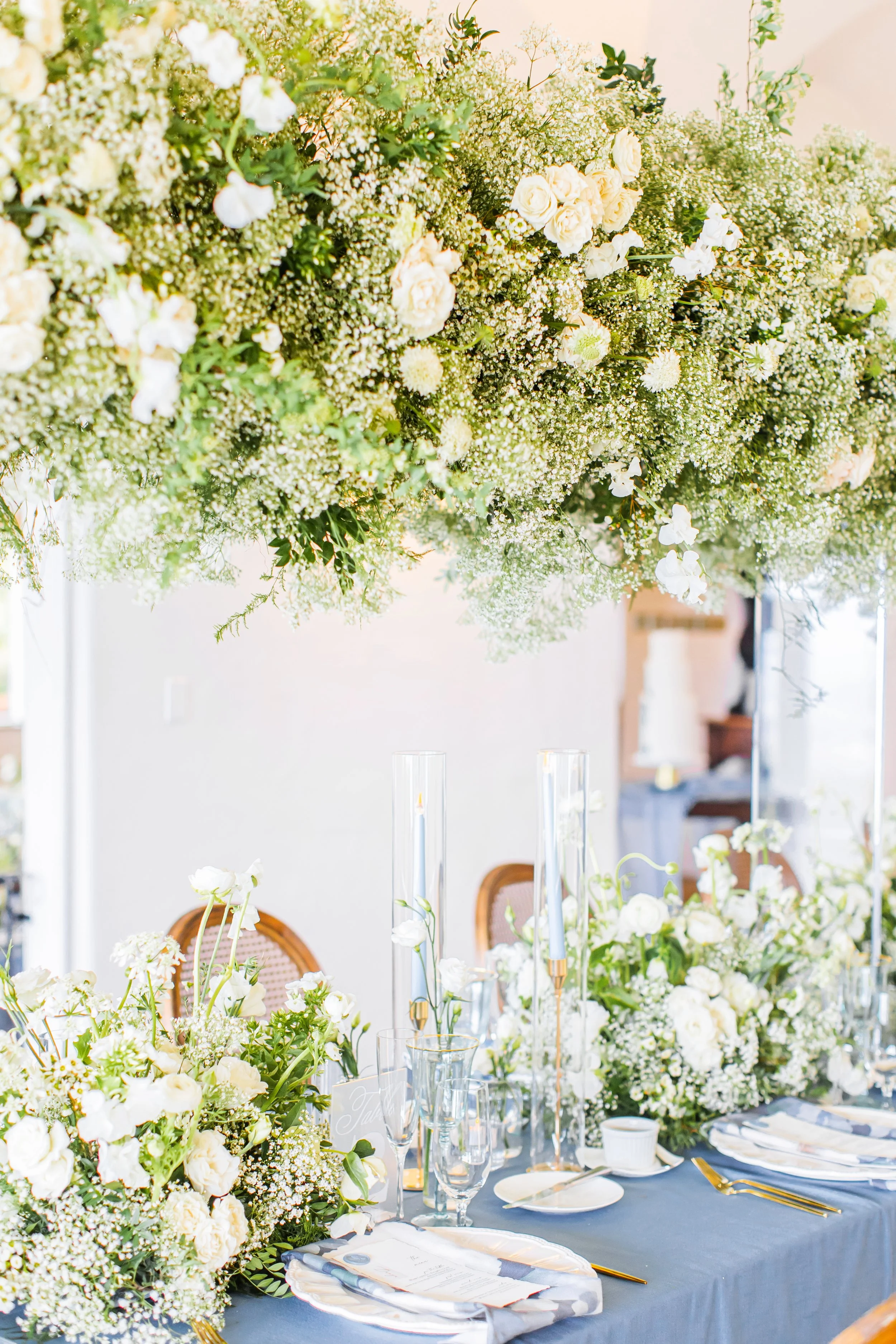 Elegant table setup with white floral centerpieces, tall glass candle holders, and place settings with gold accents. Overhead, a large, lush floral arrangement of white roses, baby's breath, and greenery.