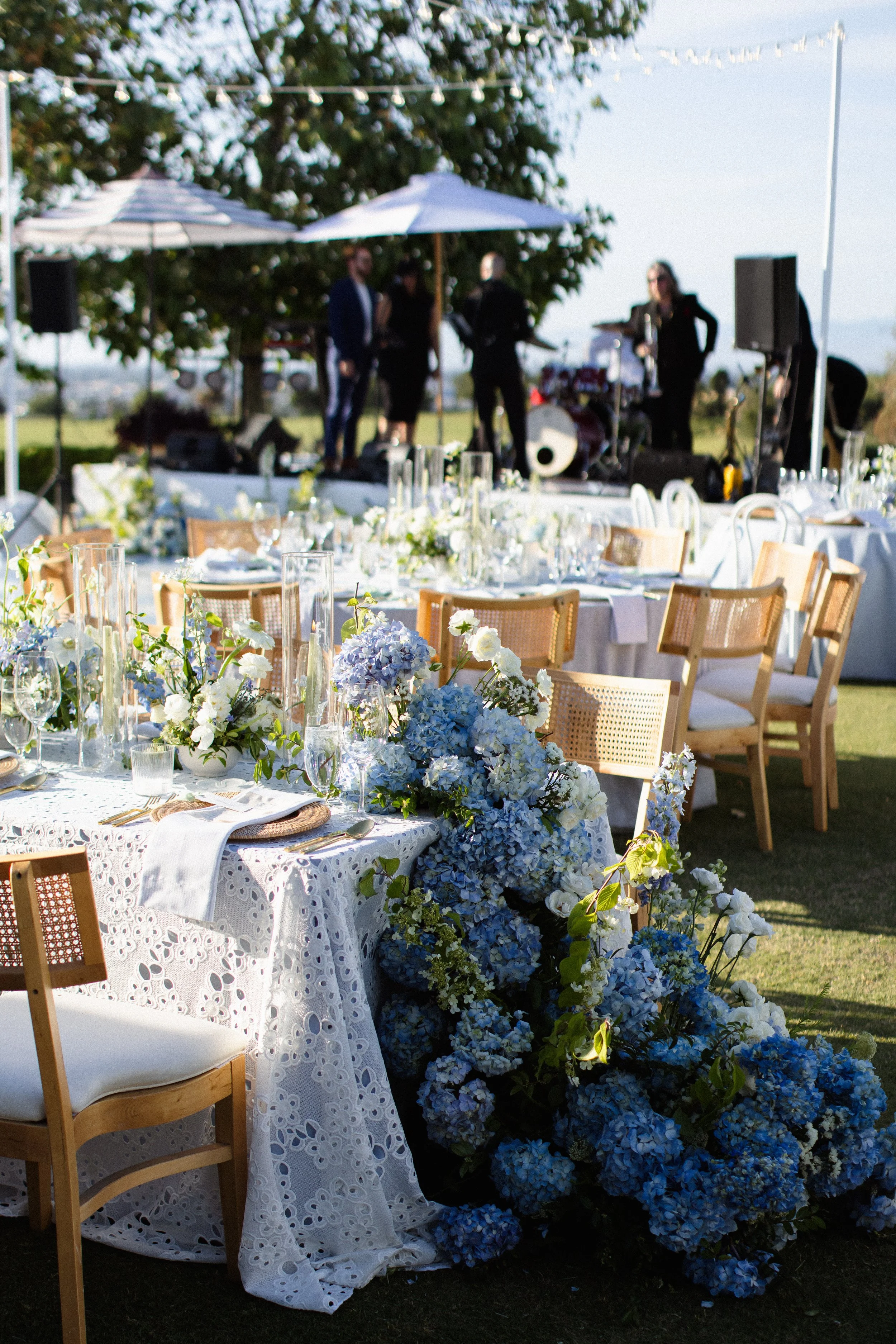 Elegant outdoor event setup with framed tables adorned with white lace tablecloths, blue and white floral centerpieces, and glassware, with a live band performing under umbrellas in the background. A Rolling Hills Country Club wedding.