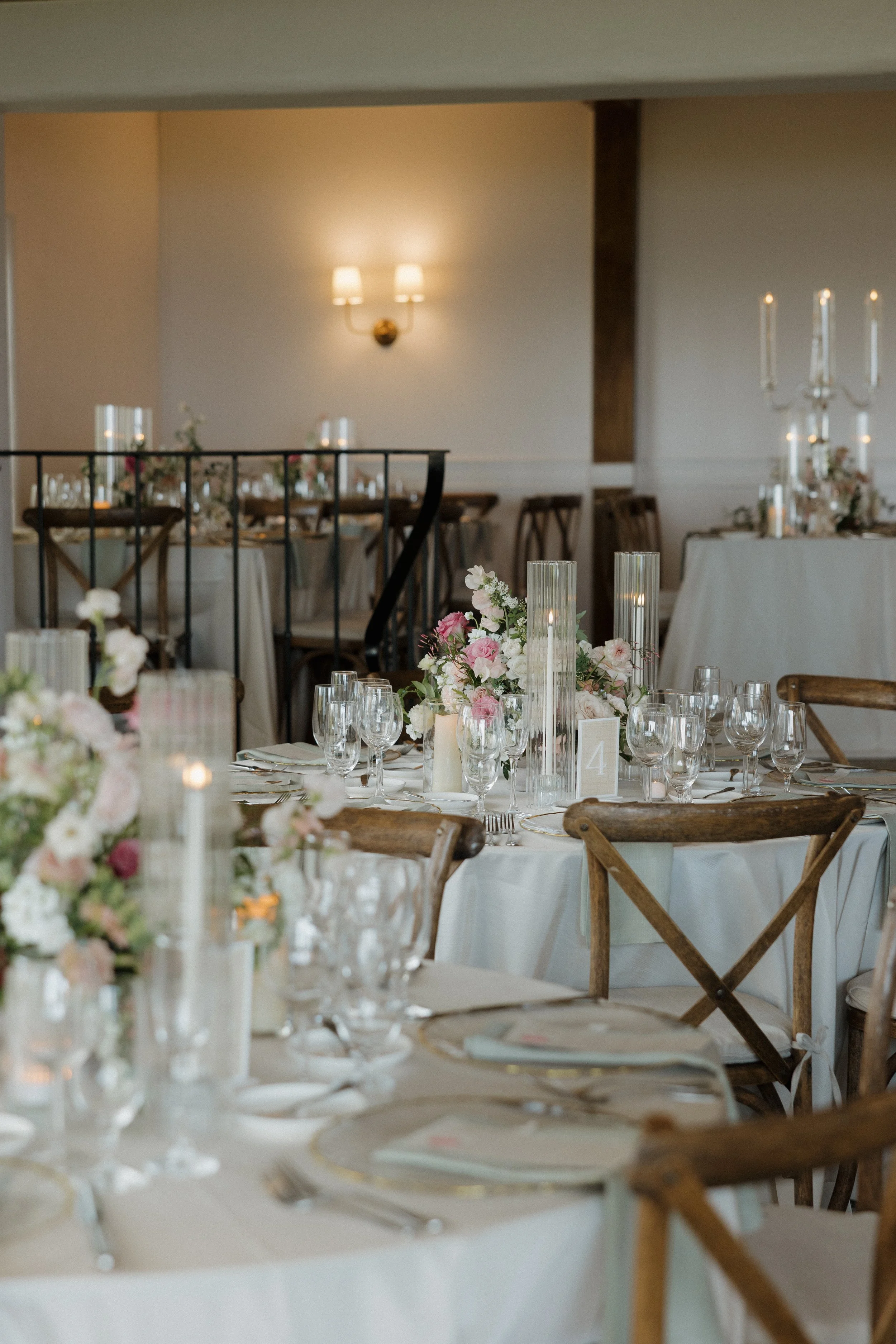 Elegant wedding reception tables with floral centerpieces, candles, glassware, and wooden chairs in a decorated hall.