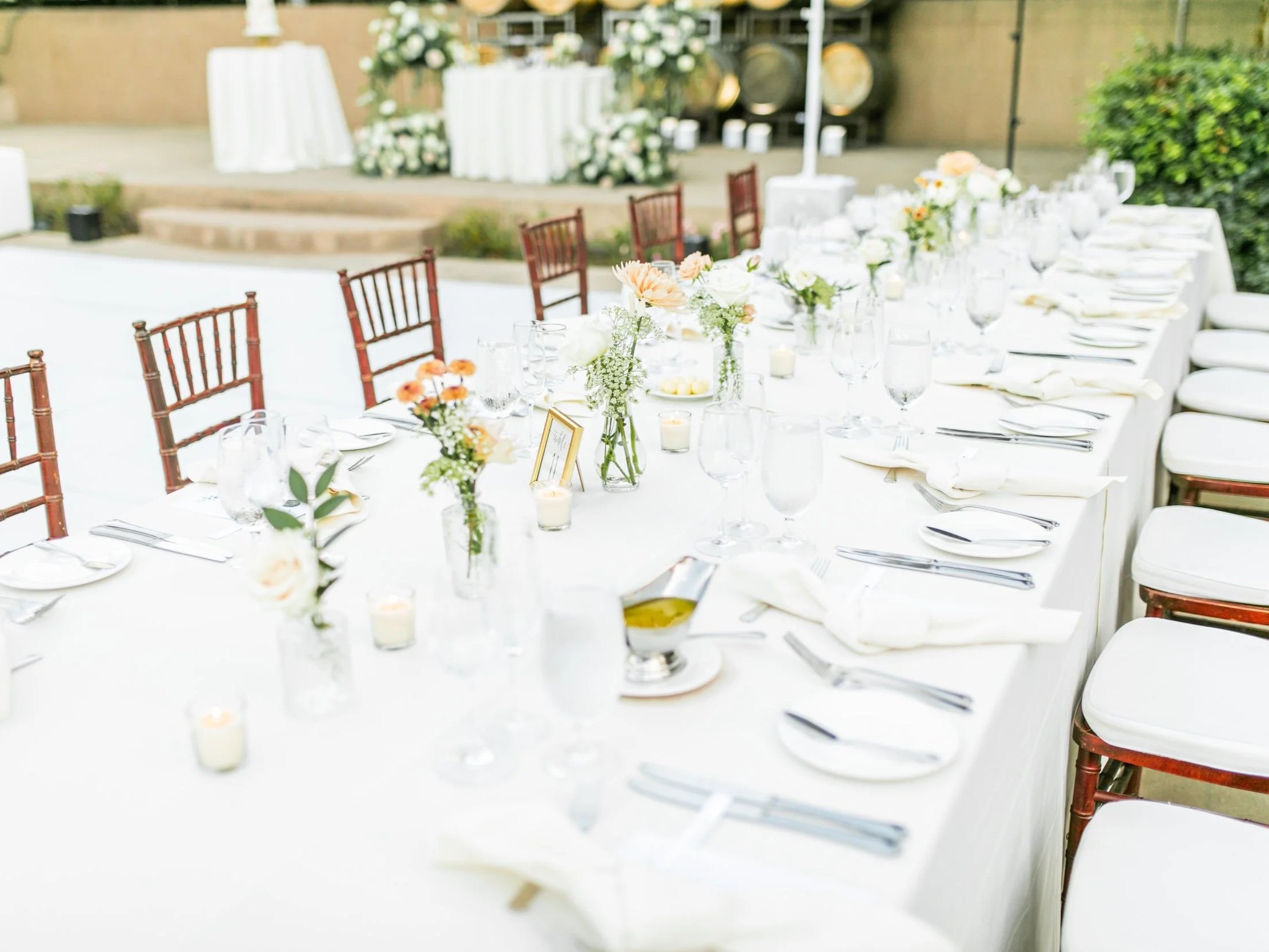 Wedding reception table decorated with white tablecloths, floral centerpieces, candles, and elegant tableware set outdoors.
