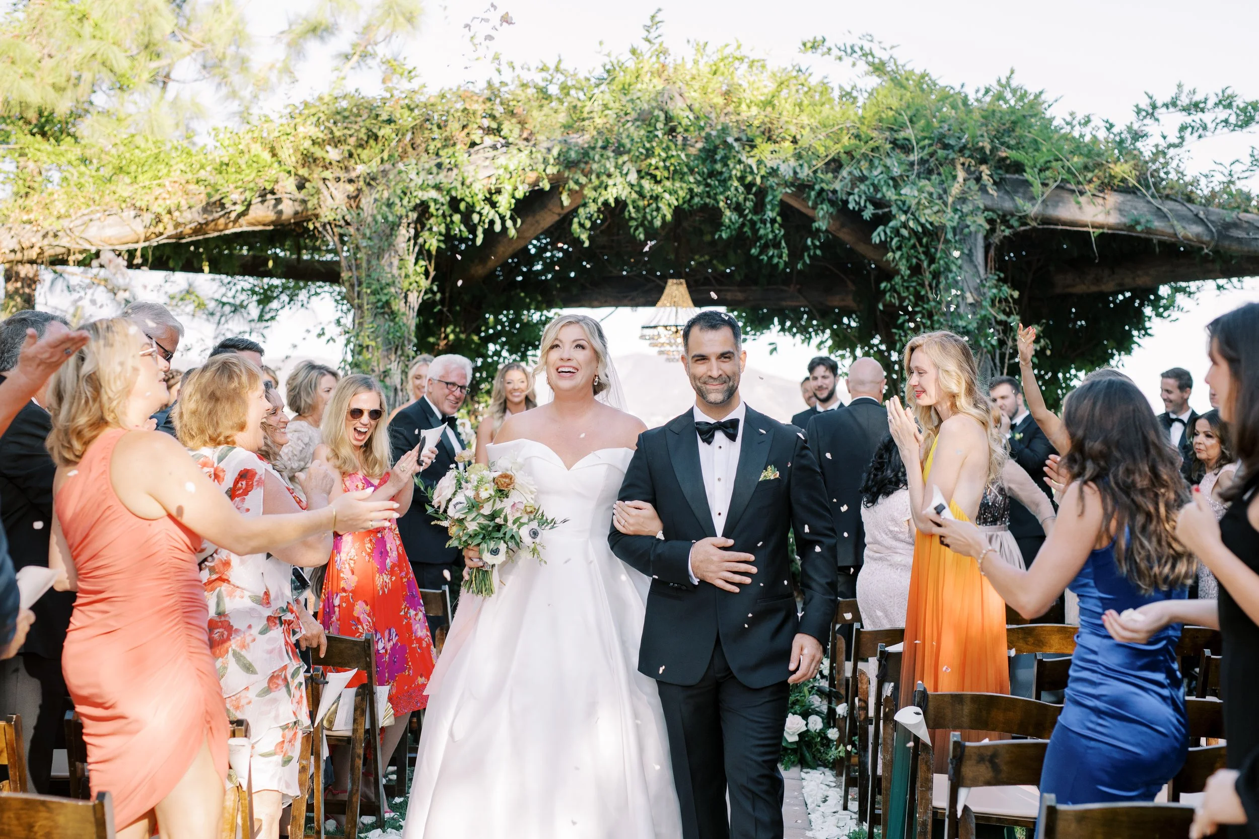 Bride and Groom walking down the aisle with Rose Arbor in the background covered in vines. Floral petals being tossed and guests standing applauding in excitement.