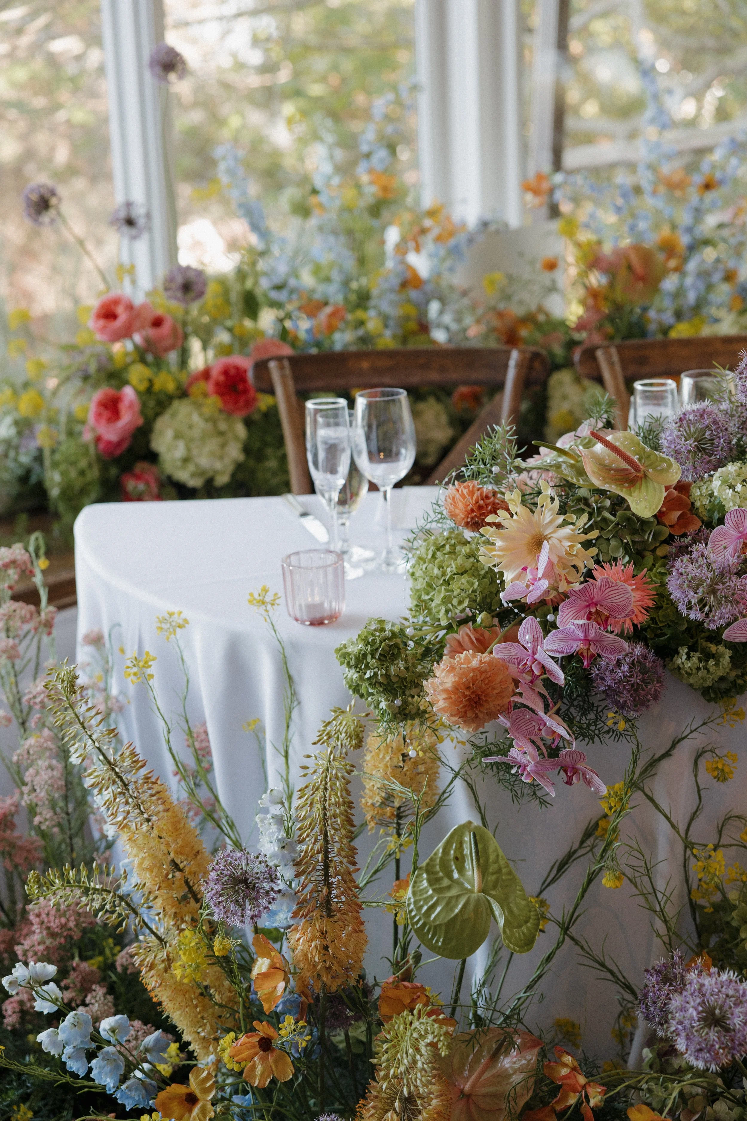 A table decorated with an abundance of colorful flowers including pink, yellow, orange, purple, and green, set with glasses and a white tablecloth in a bright room with large windows and greenery outside.