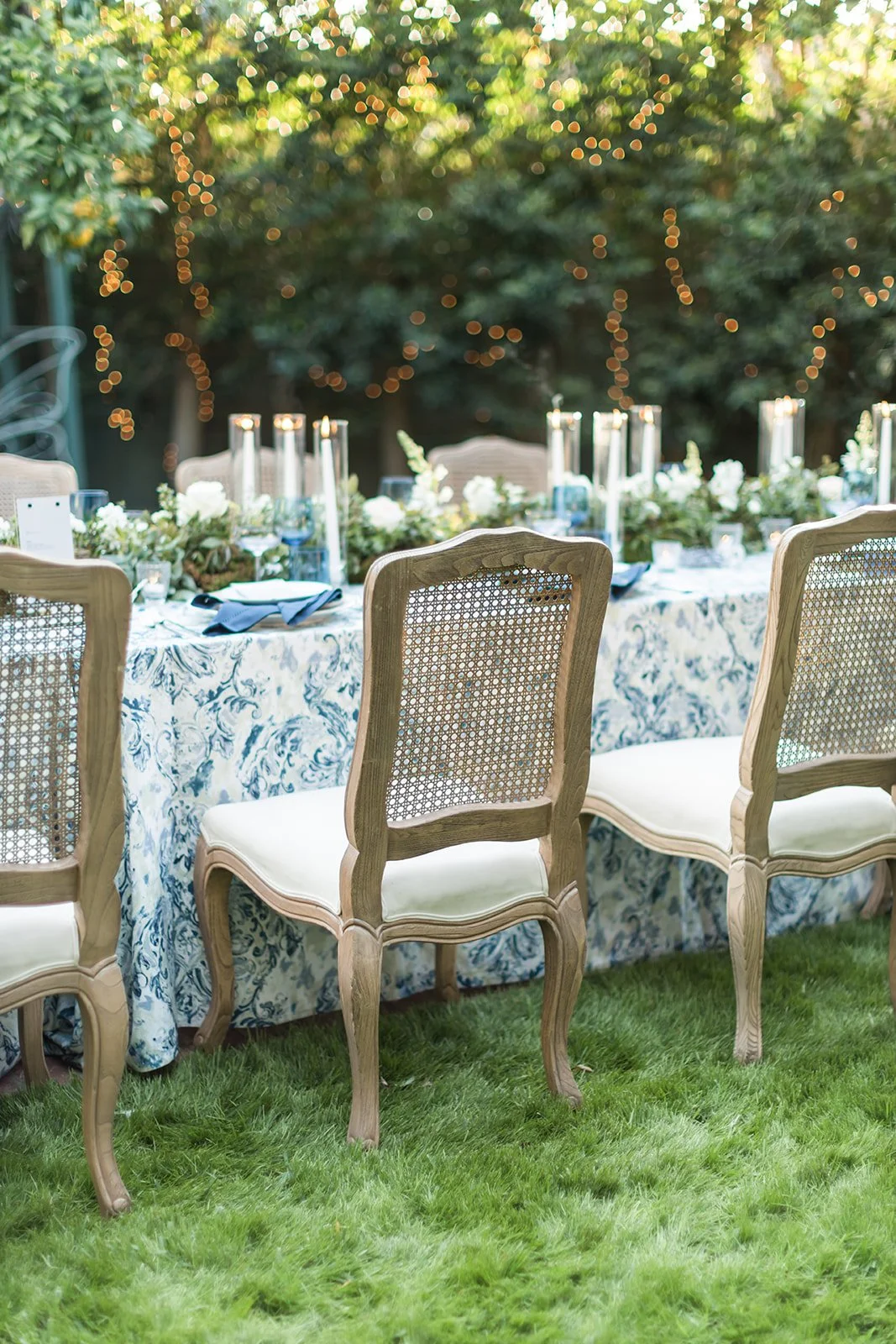 Elegant outdoor dining table set with floral tablecloth, tall candles, and glassware, surrounded by vintage-style wooden chairs on a grassy lawn, with string lights and trees in the background.