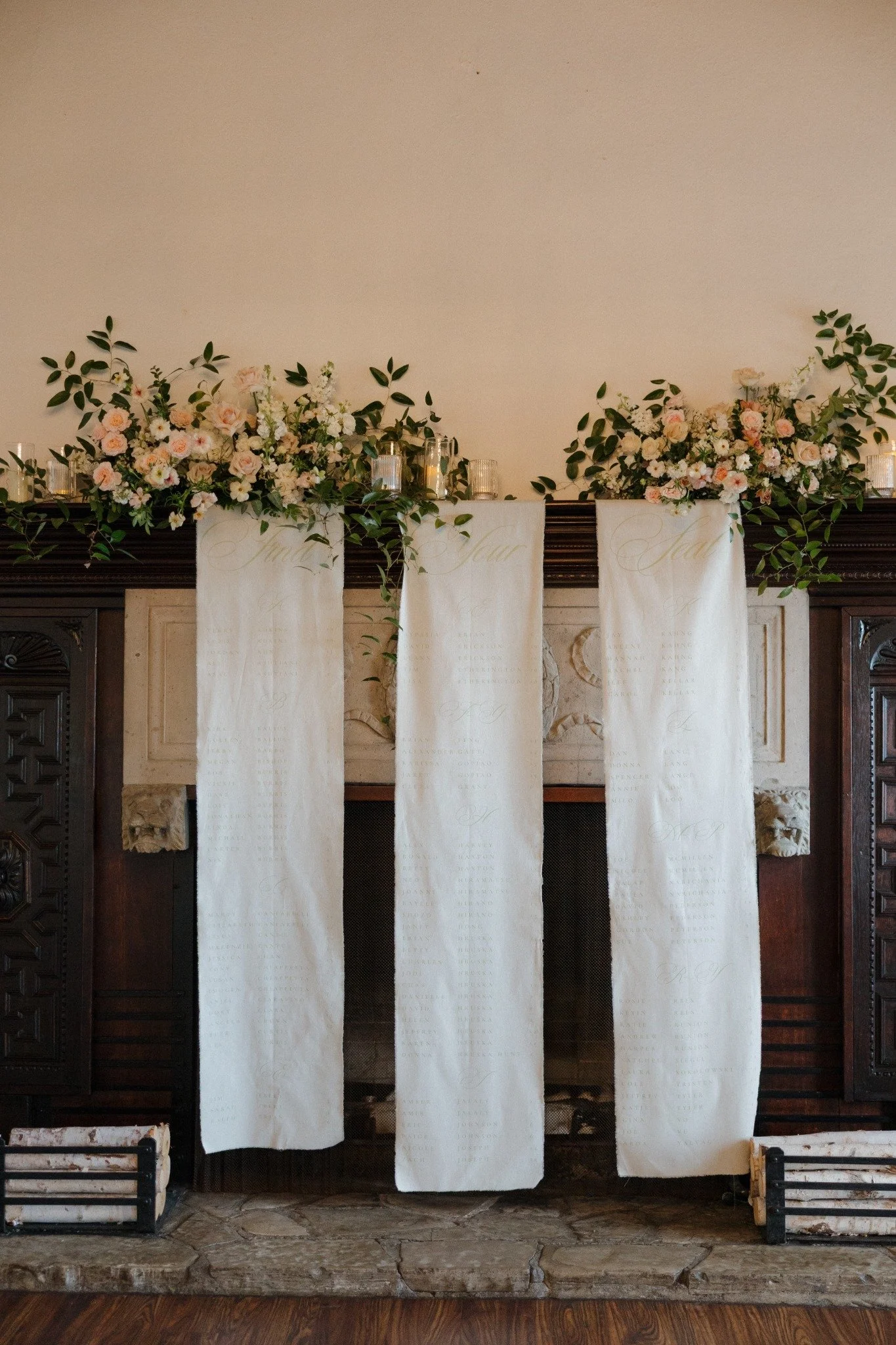 Elegant fireplace decorated with three white fabric panels with cursive writing, topped with floral arrangements of pink and white roses and greenery, with glass candle holders and candles on the mantle. Seating Chart display.