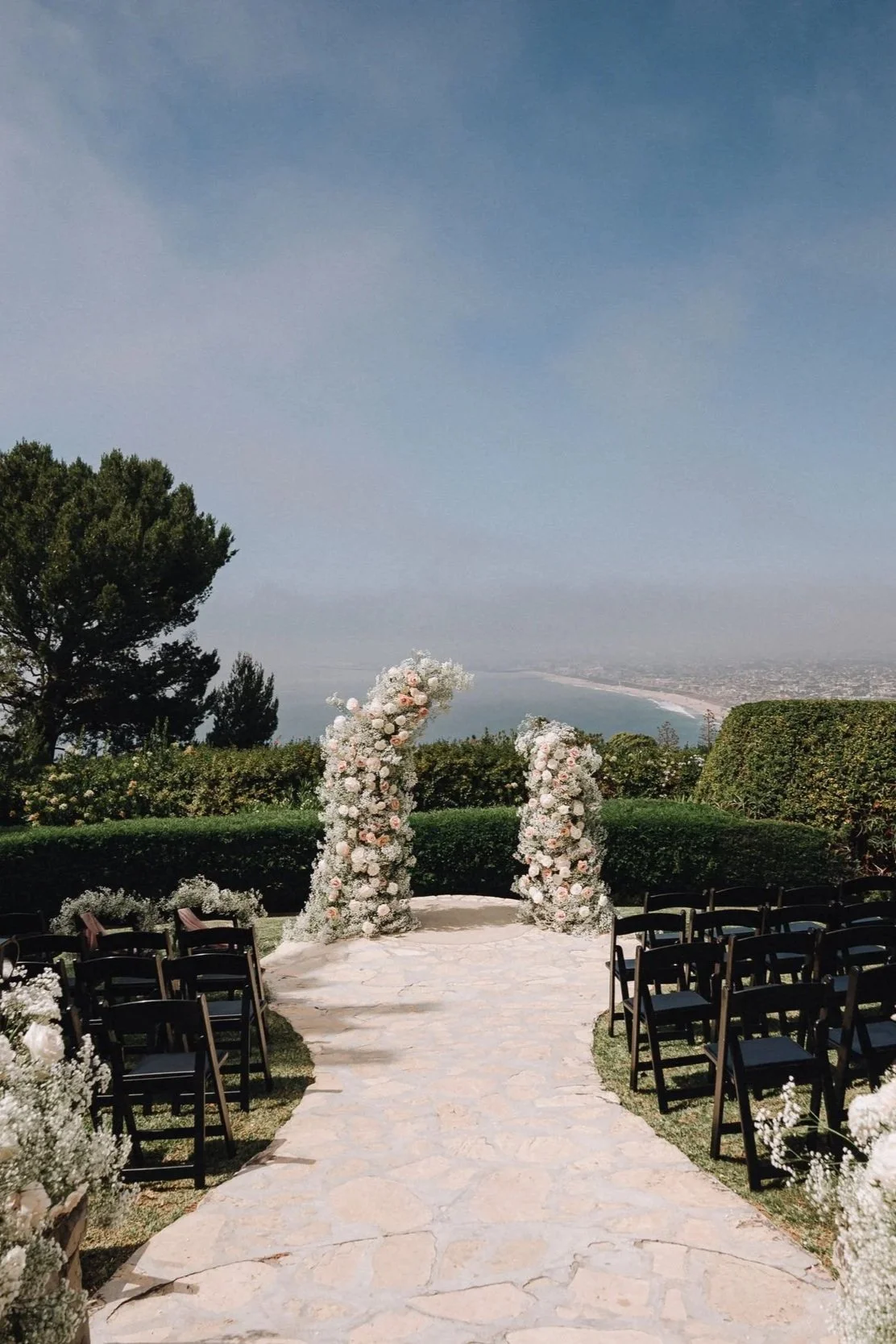 Outdoor wedding ceremony setup on a stone aisle with floral arches and black chairs, overlooking a landscape with trees, a body of water, and a cloudy sky. A La Venta Inn Wedding.
