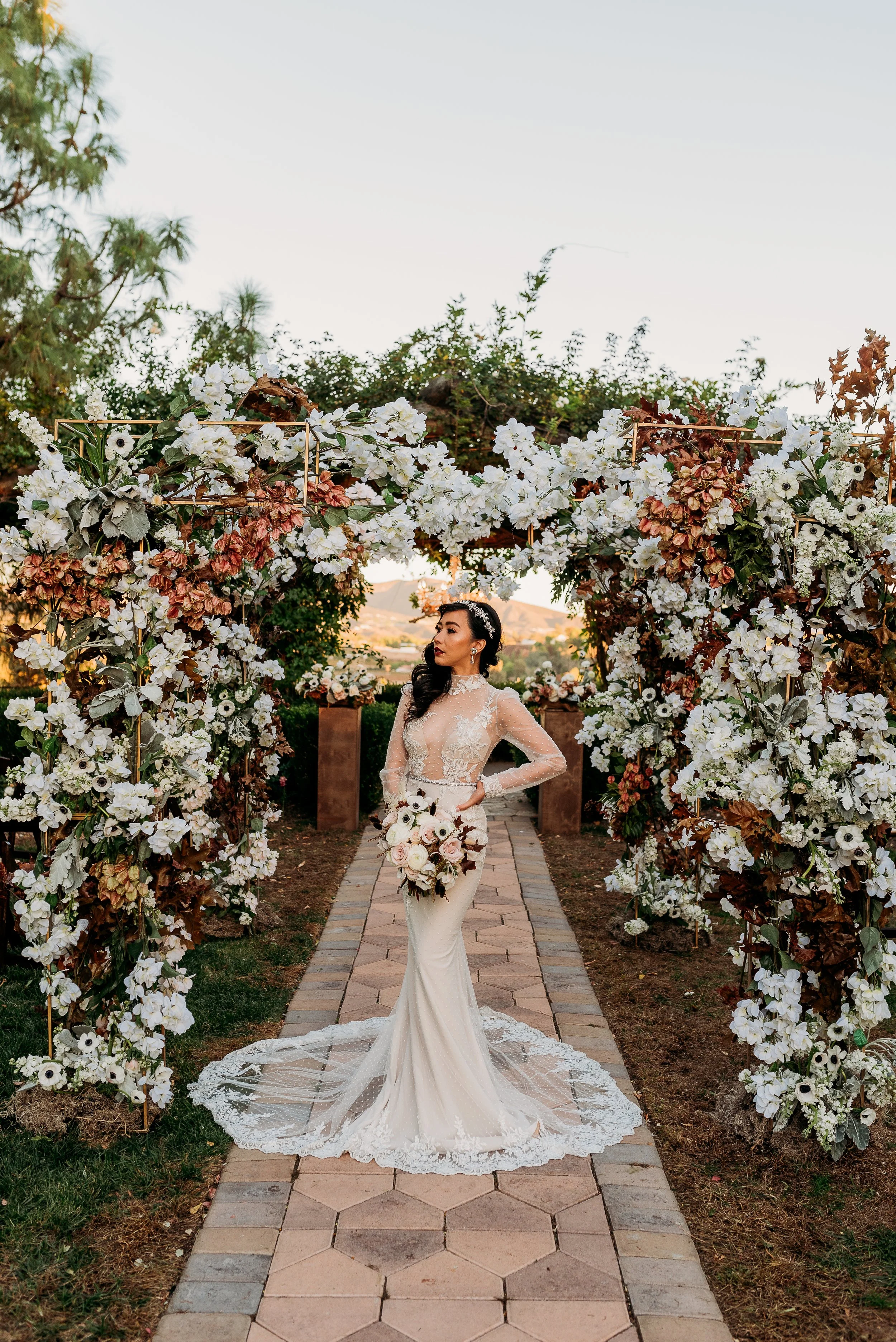 Bride portrait under floral ceremony arch at South Coast Winery wedding in Temecula featuring white blooms, autumn tones, and elegant lace gown