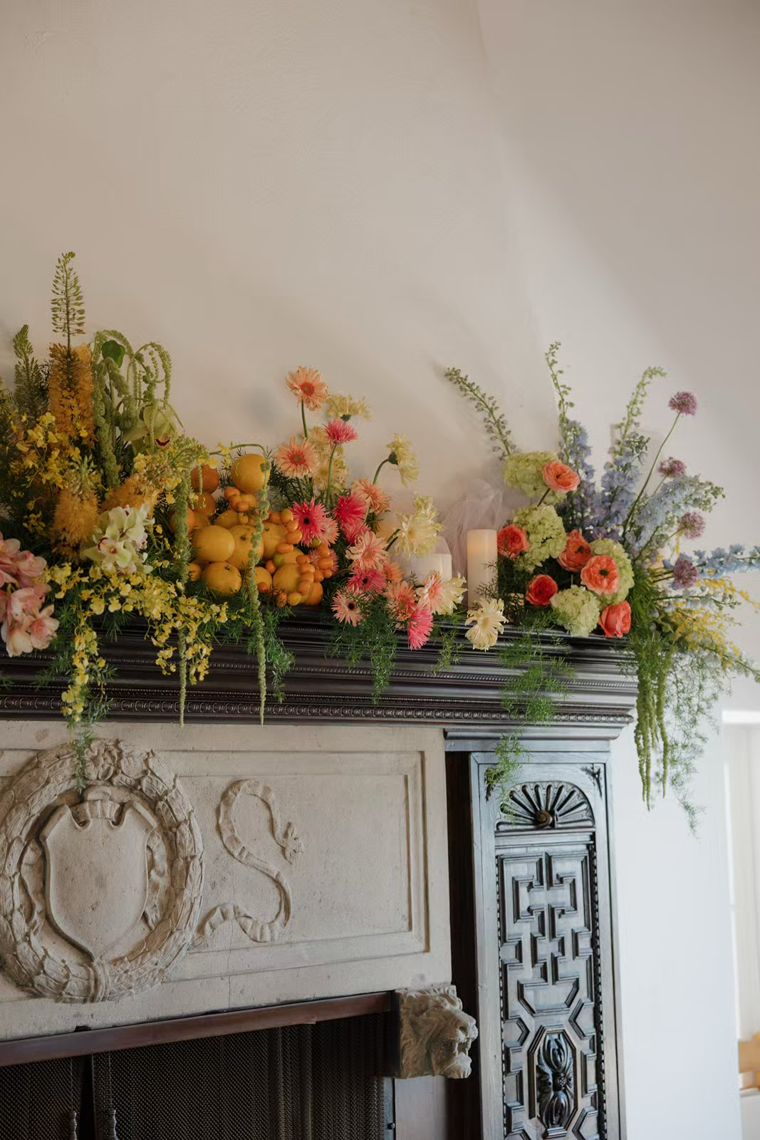 the top of a fire place with large bold colored flowers, and fruit arrangements 