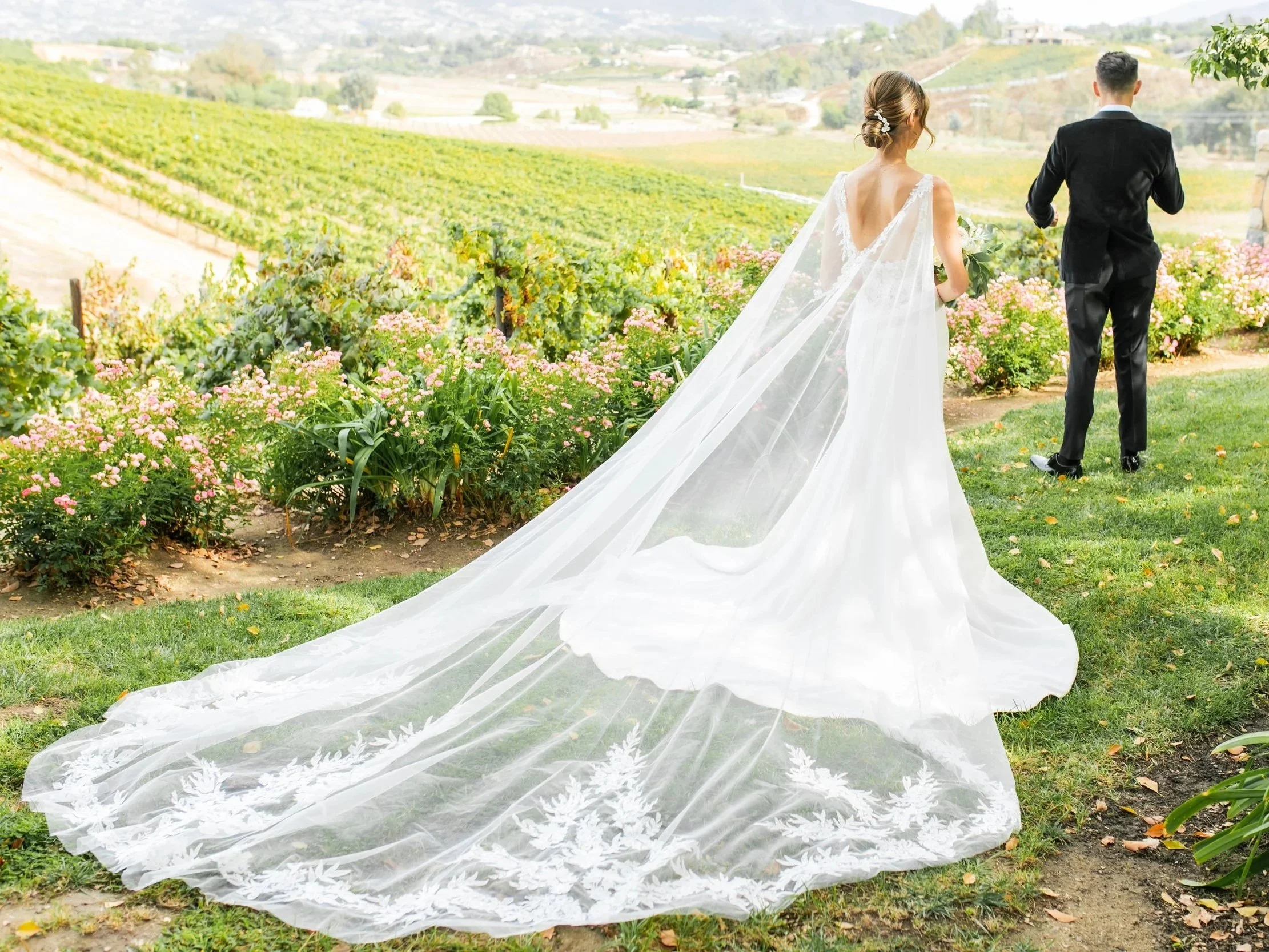 A bride and groom walking away from camera in a scenic vineyard during daytime. The bride wears a white wedding gown with a long, flowing train and veil, and holds a bouquet. The groom is dressed in a dark suit. Green grass and pink flowers border the pathway, with rolling hills and vineyards in the background.