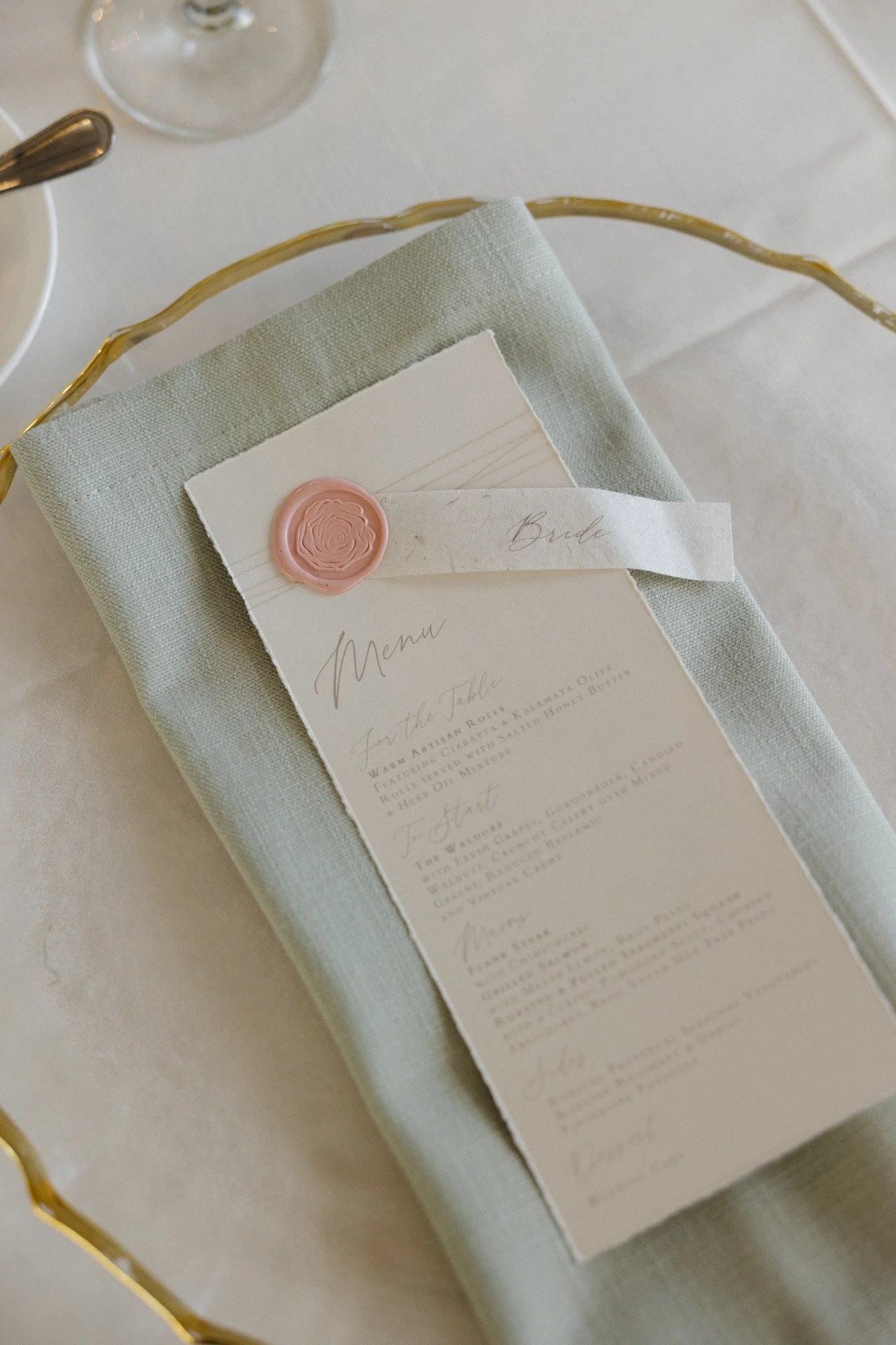 Wedding menu with a pink wax seal and a 'Bride' label on a folded napkin, placed on a table with a white tablecloth, glassware, and silverware.
