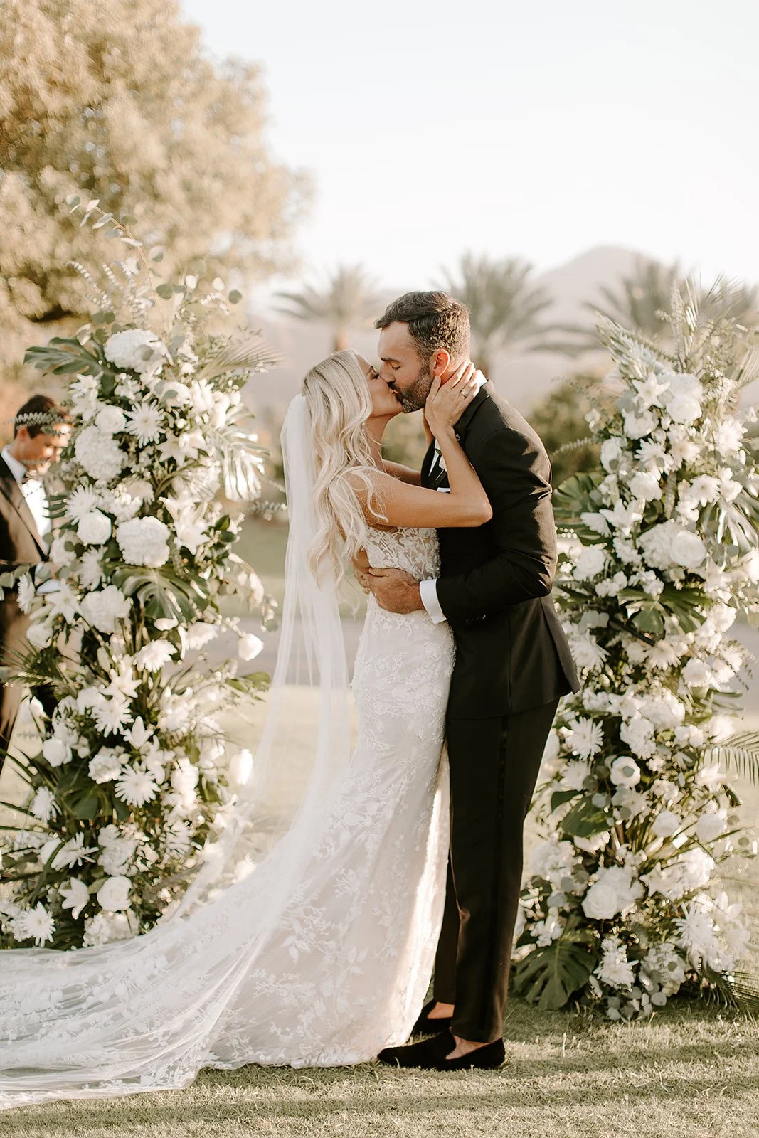 A bride and groom kiss during their outdoor wedding ceremony, surrounded by large floral arrangements with white flowers and greenery. An Indian Wells Golf Resort Wedding.