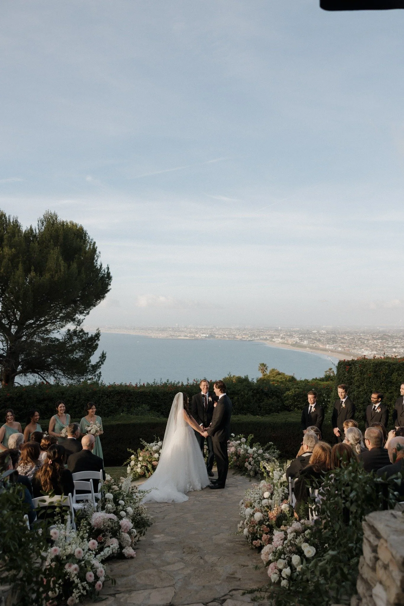 A wedding ceremony outdoors overlooking a city and body of water, with a bride and groom holding hands and facing each other, surrounded by guests and bridesmaids and groomsmen, with floral decorations on the aisle and in front of the couple.
