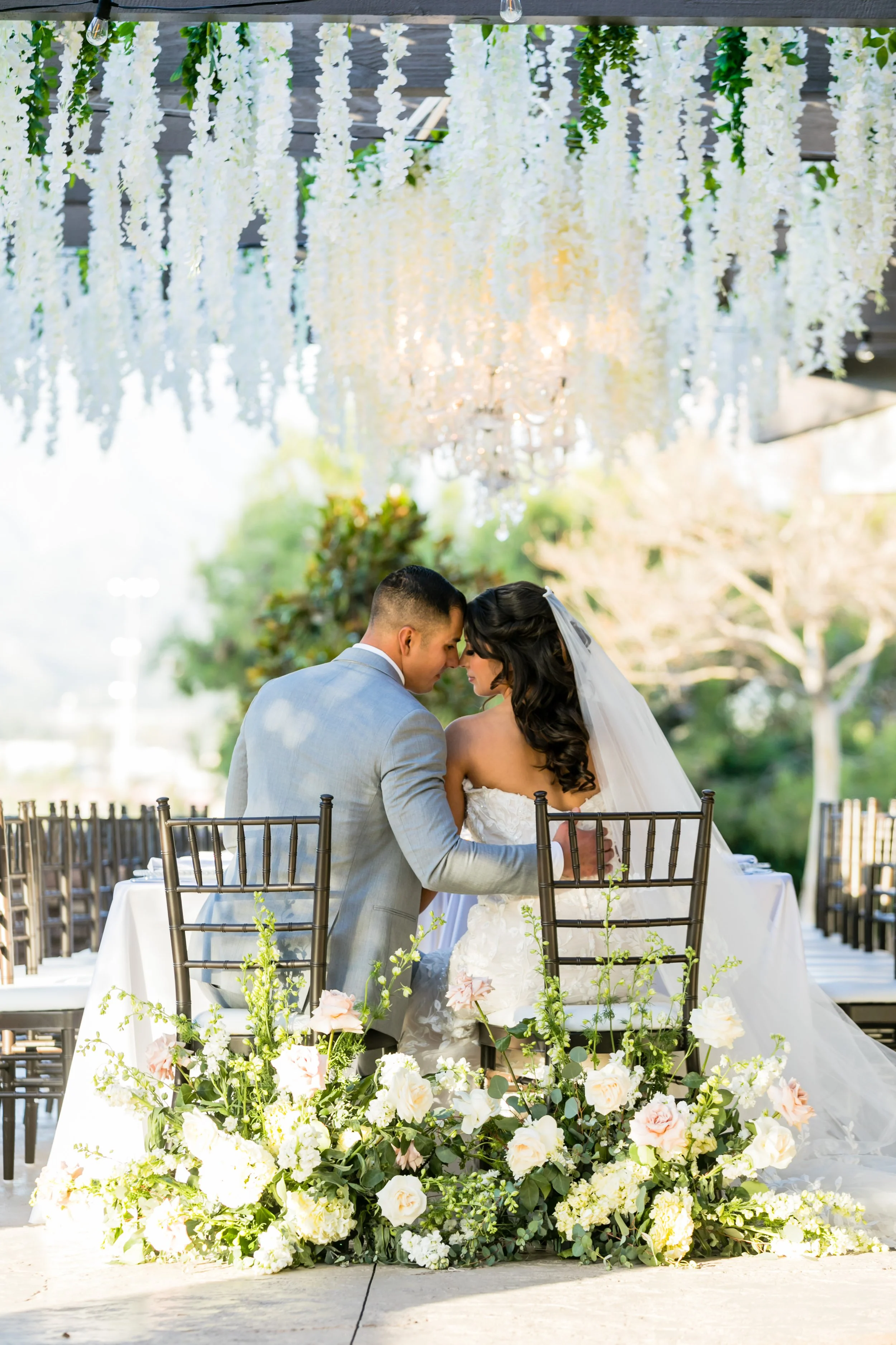 A bride and groom sitting closely together at a decorated outdoor wedding reception. The scene features floral arrangements at the forefront, hanging white floral decorations above, and lush green trees in the background.