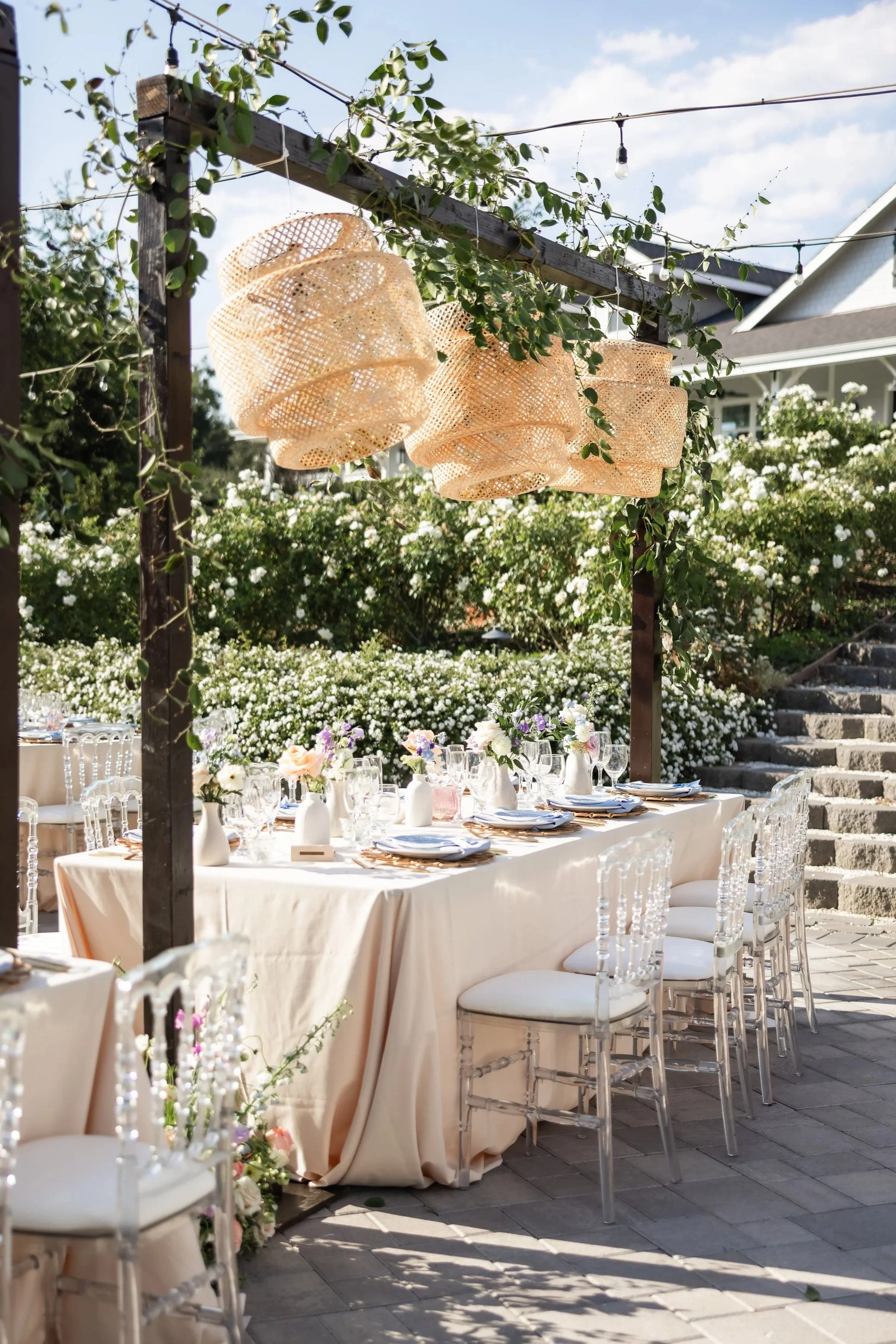 Outdoor wedding or event setup with a long table decorated with white tablecloth, floral centerpieces, and transparent chairs. Overhead, woven lanterns are suspended on a wooden beam, with greenery and white flowering bushes in the background, and a house with stairs visible.