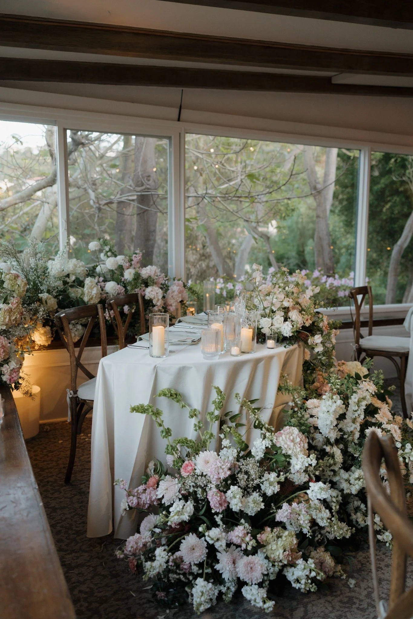 A decorated table at a wedding reception with white tablecloth, surrounded by pink and white floral arrangements, candles, and chairs, with a large window showing trees outside.