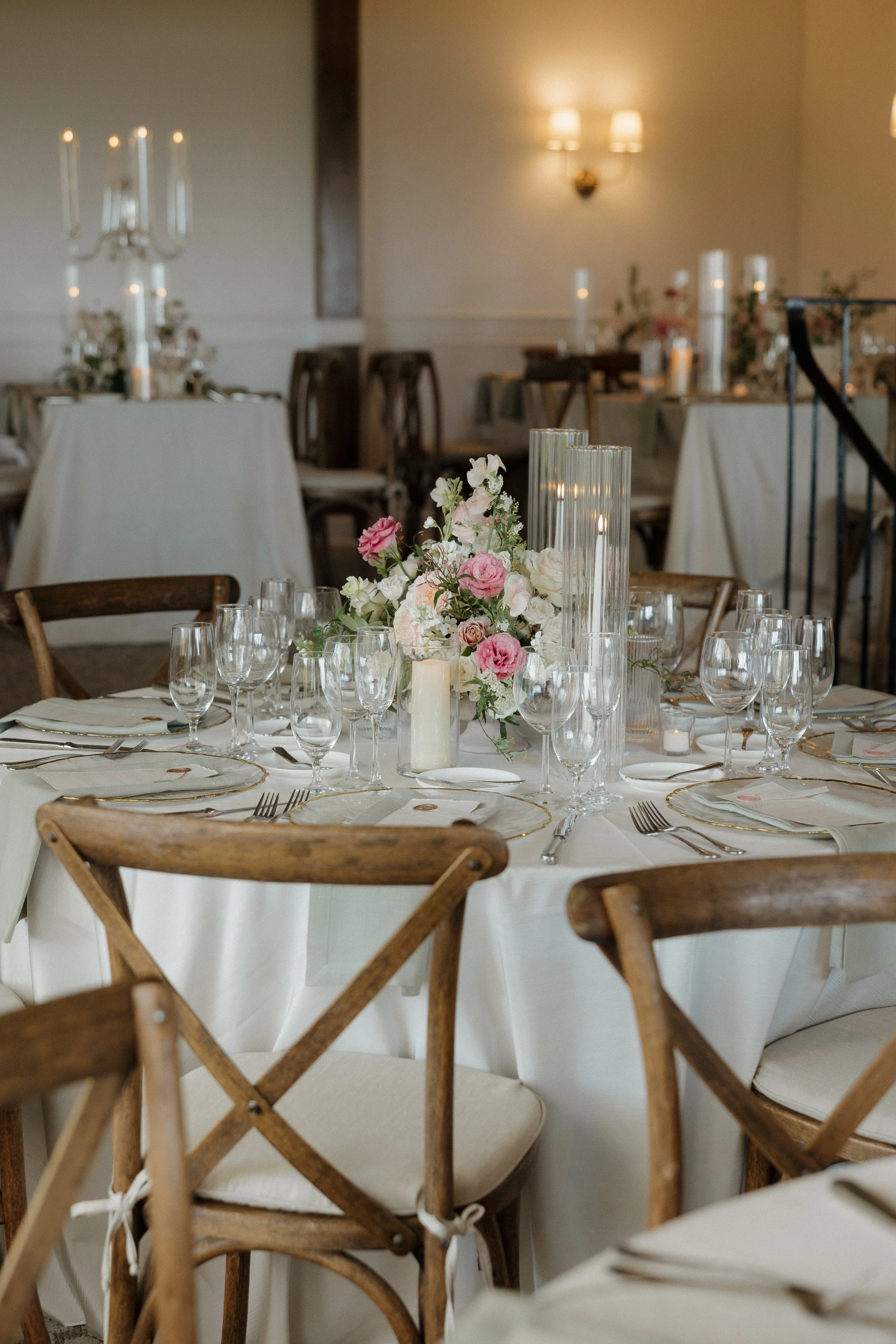 Elegant banquet table decorated with pink and white flowers, glass candles, and tall candles in a softly lit room with additional decorated tables in the background.