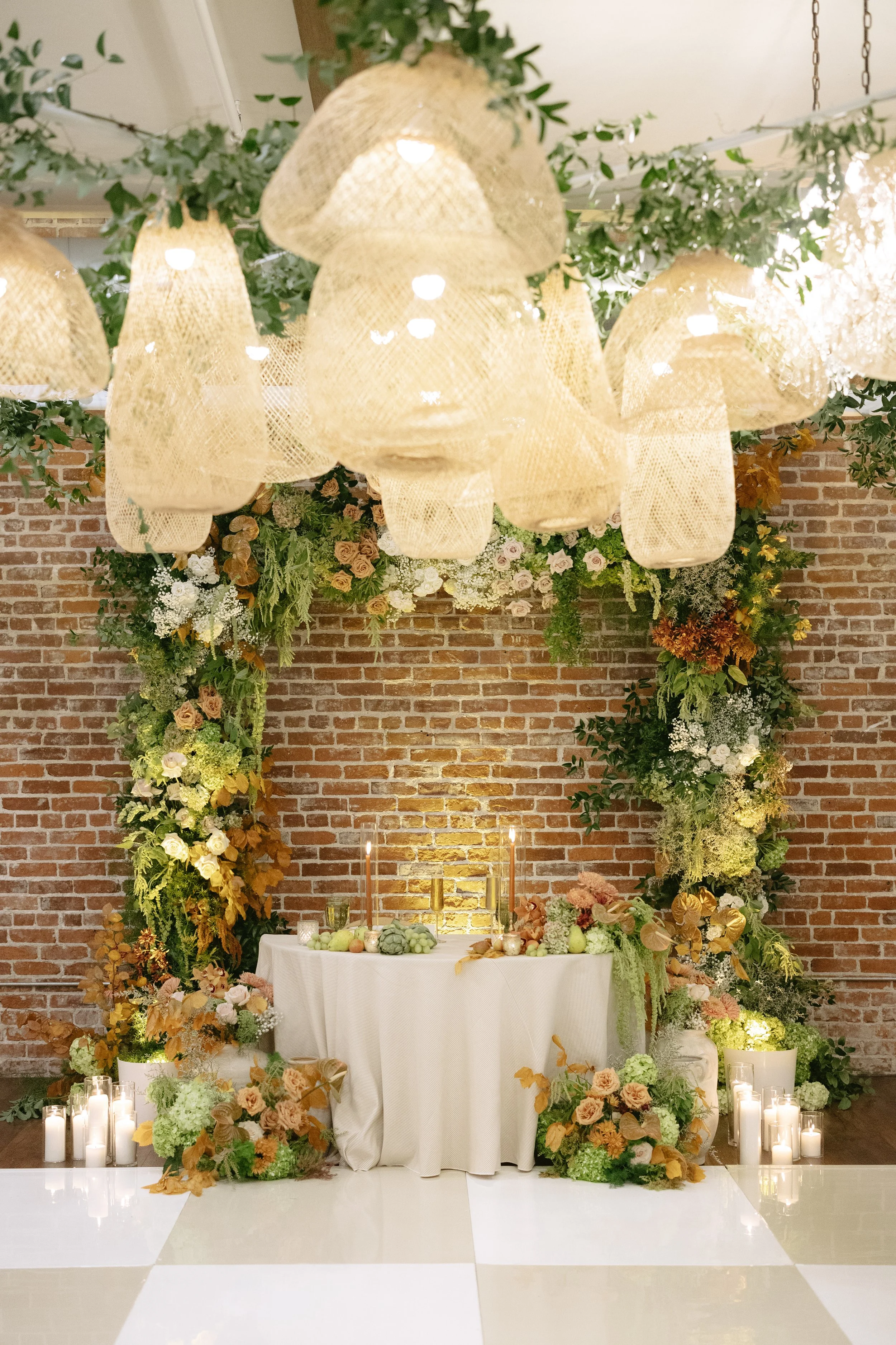 Decorated wedding or event altar featuring a floral arch, candles, and a table with fruit and flowers, set against a brick wall. Franciscan Gardens wedding in San Juan Capistrano.