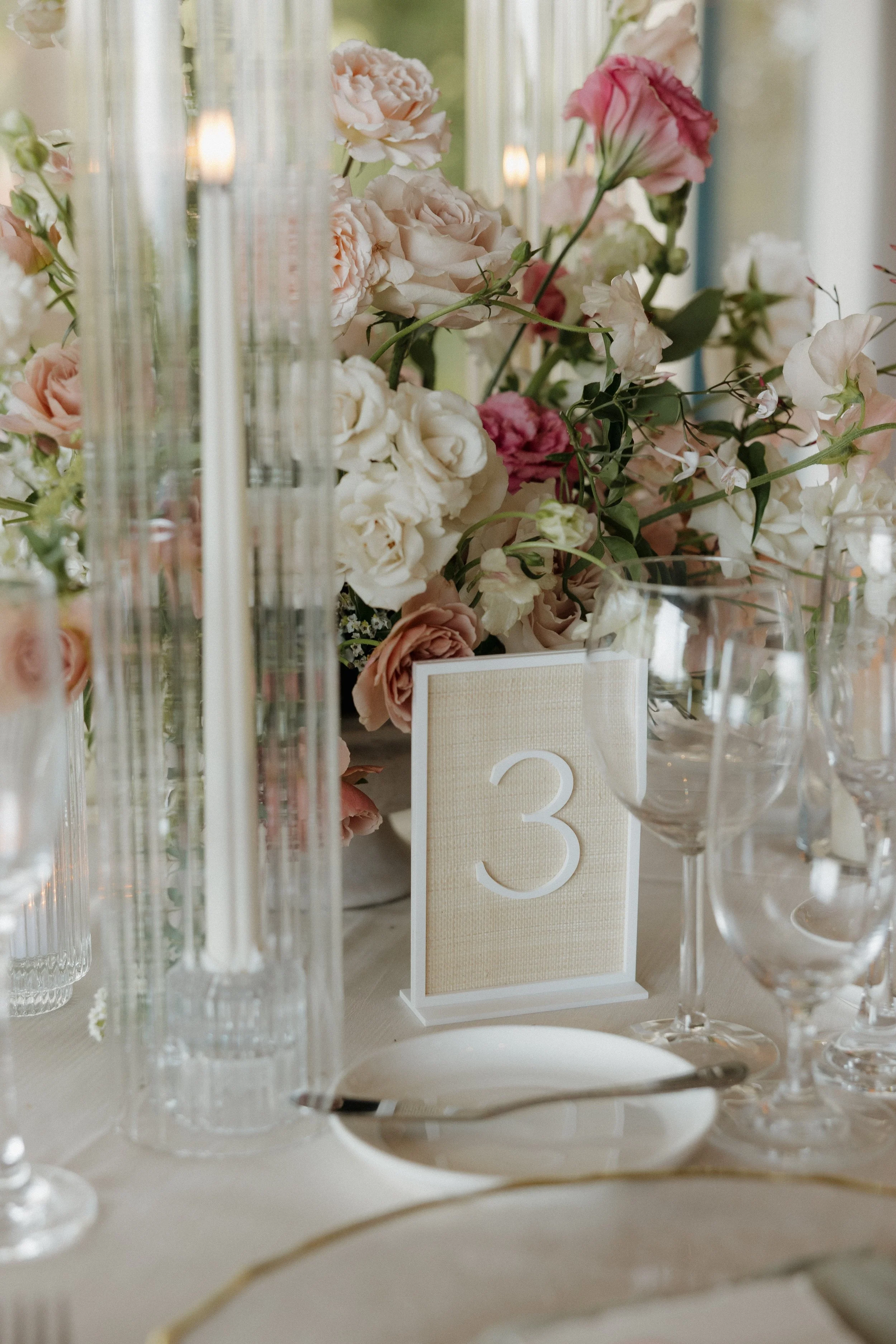 Elegant table setting with pink and white floral centerpiece, tall glass candle holders, and a table number '3' on a beige card.