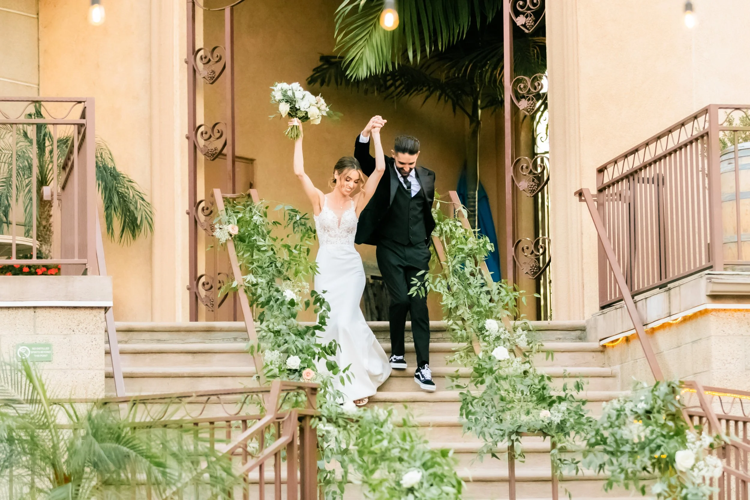 Bride and groom are holding hands and walking down the stairs with greenery and white flowers, celebrating their wedding.