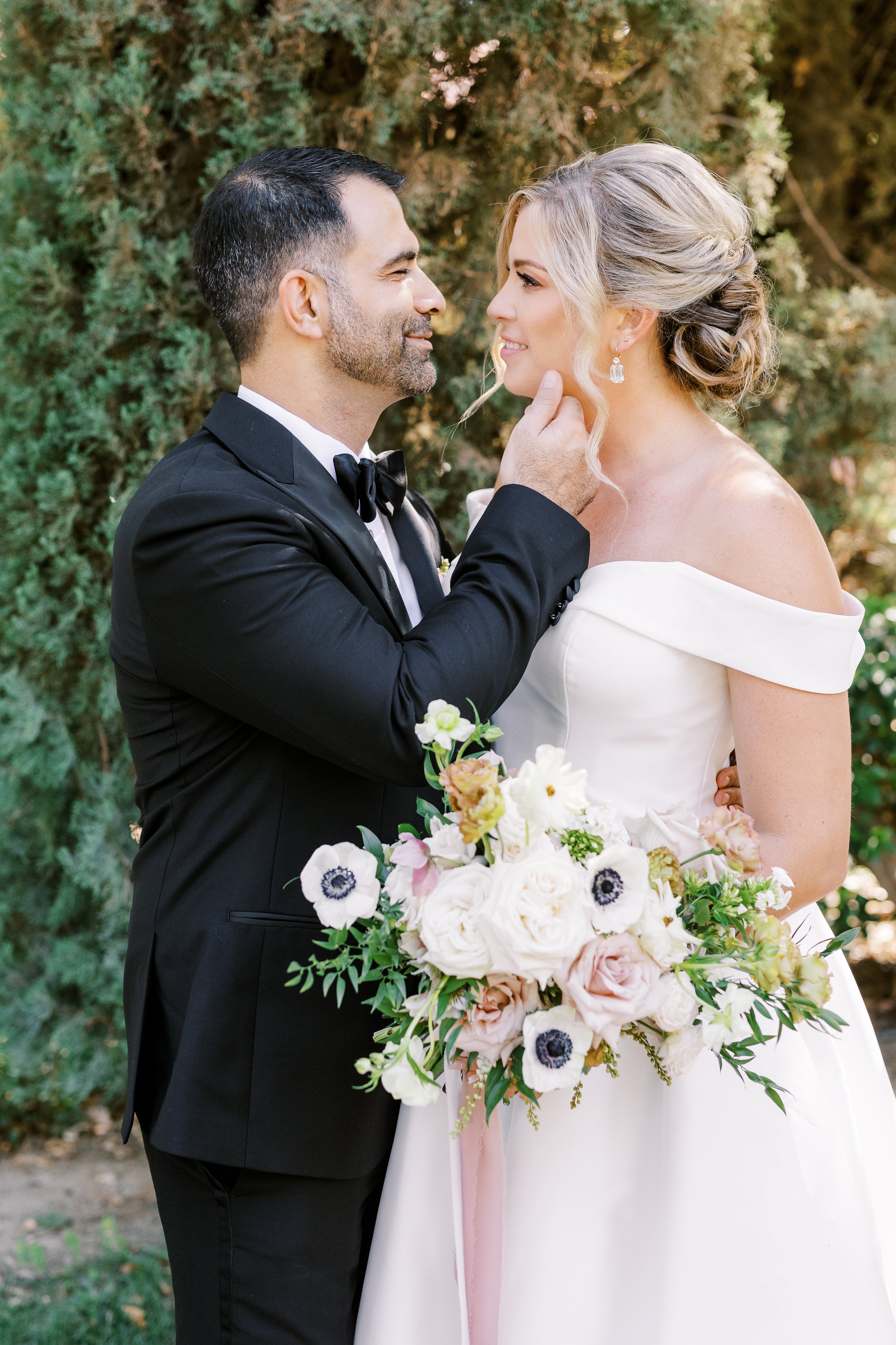 bride and groom look longingly into each other eyes on their wedding day while she is dressed in a beautiful white dress, and he is clothed in in black formal tux
