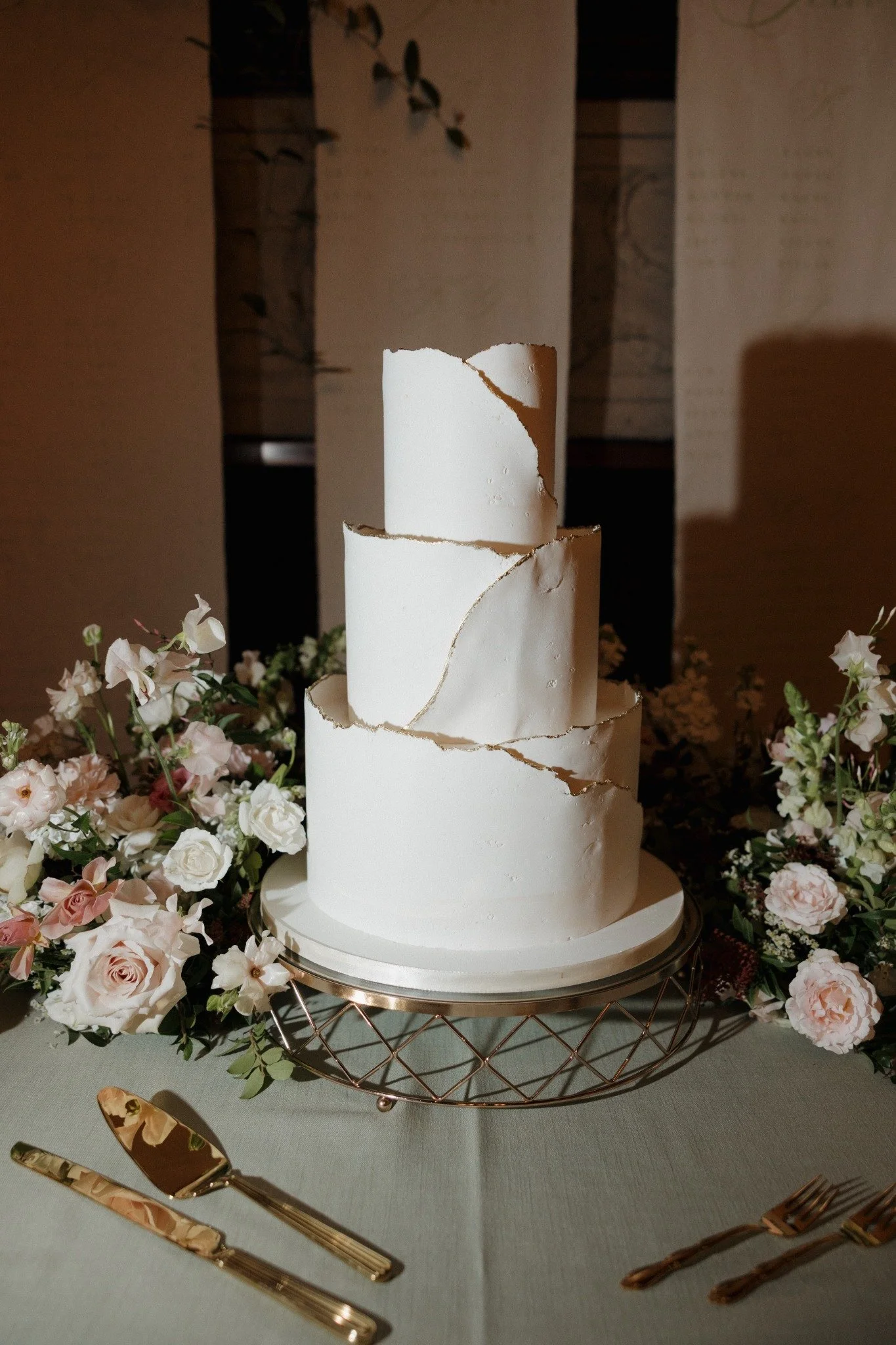 A three-tiered white wedding cake with cracked icing, surrounded by pink and white flowers on a table.