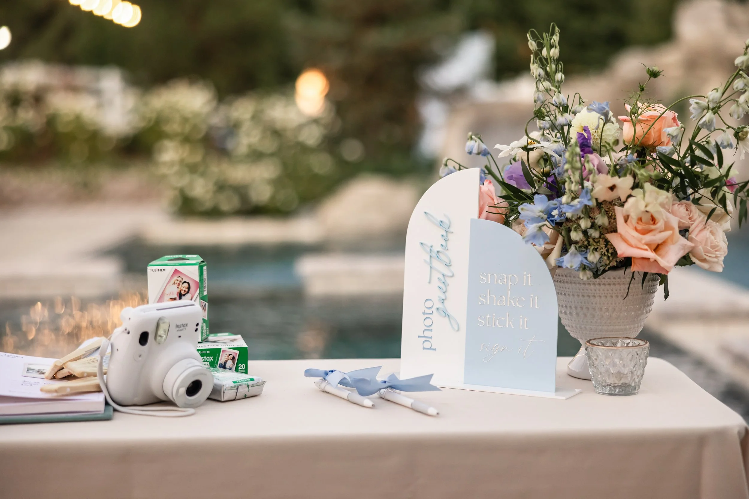 A table with a large floral arrangement, a sign that reads 'photo booth' with instructions 'snap it, shake it, stick it, sign it,' a vintage Instant camera, a box of instant film, and two pens with blue ribbons, set outdoors with blurred background