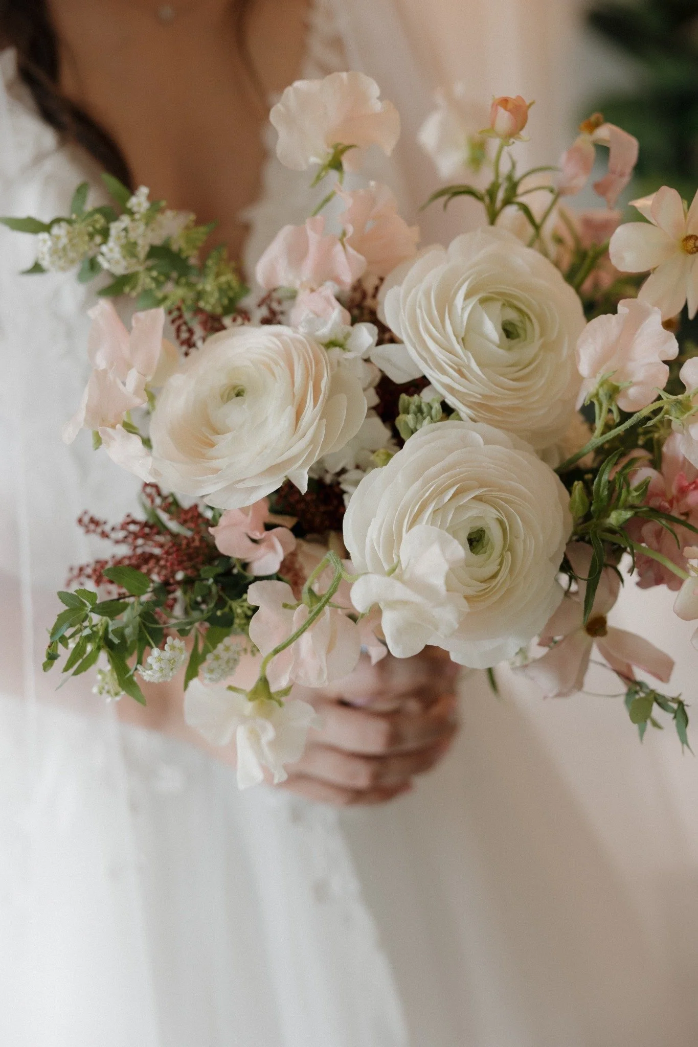A person holding a bouquet of white and light pink flowers, including ranunculus and sweet peas, with green foliage.