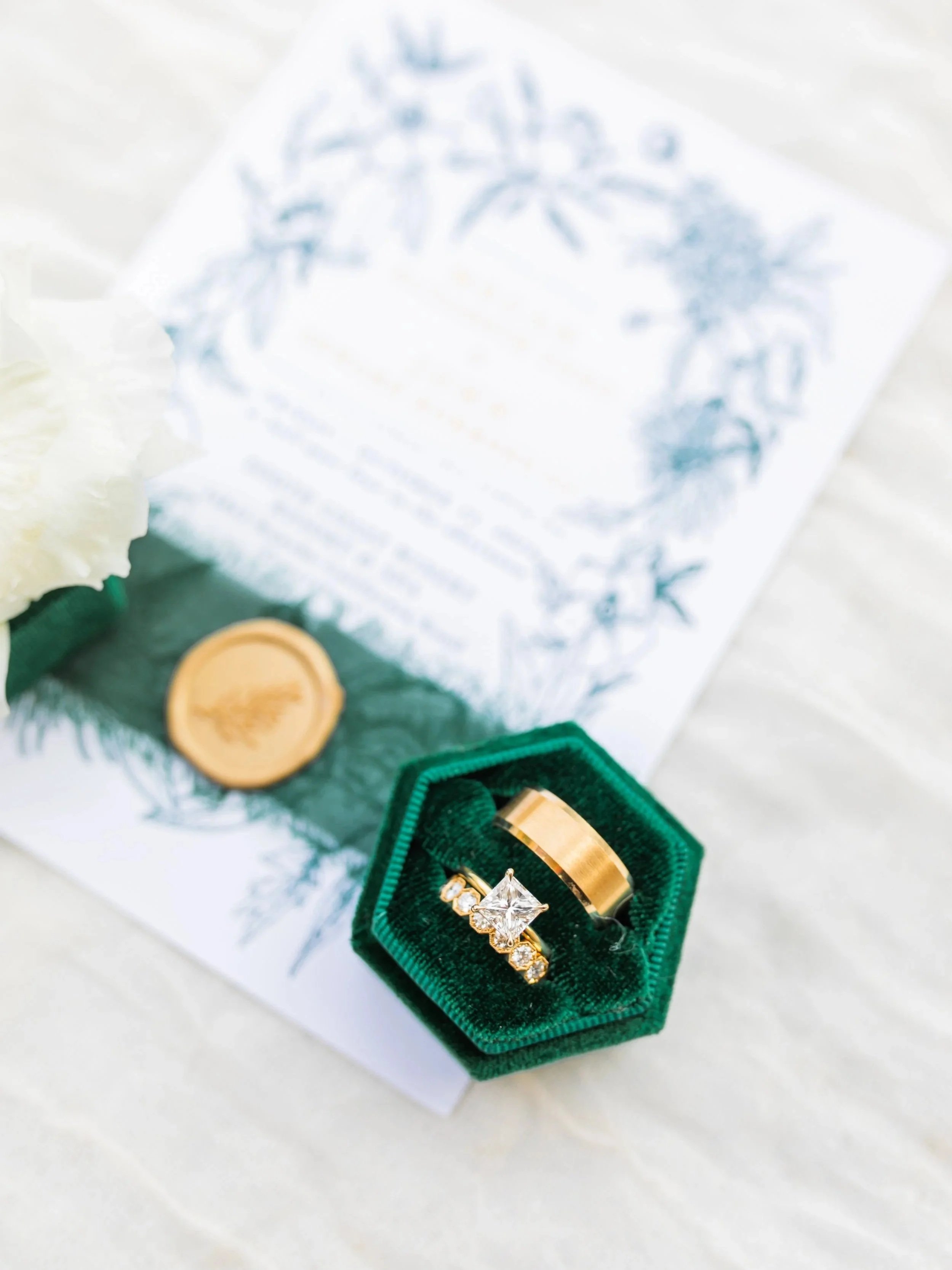 Gold wedding band and diamond engagement ring in green velvet ring box, with a wedding invitation, wax seal, and white flower in background.