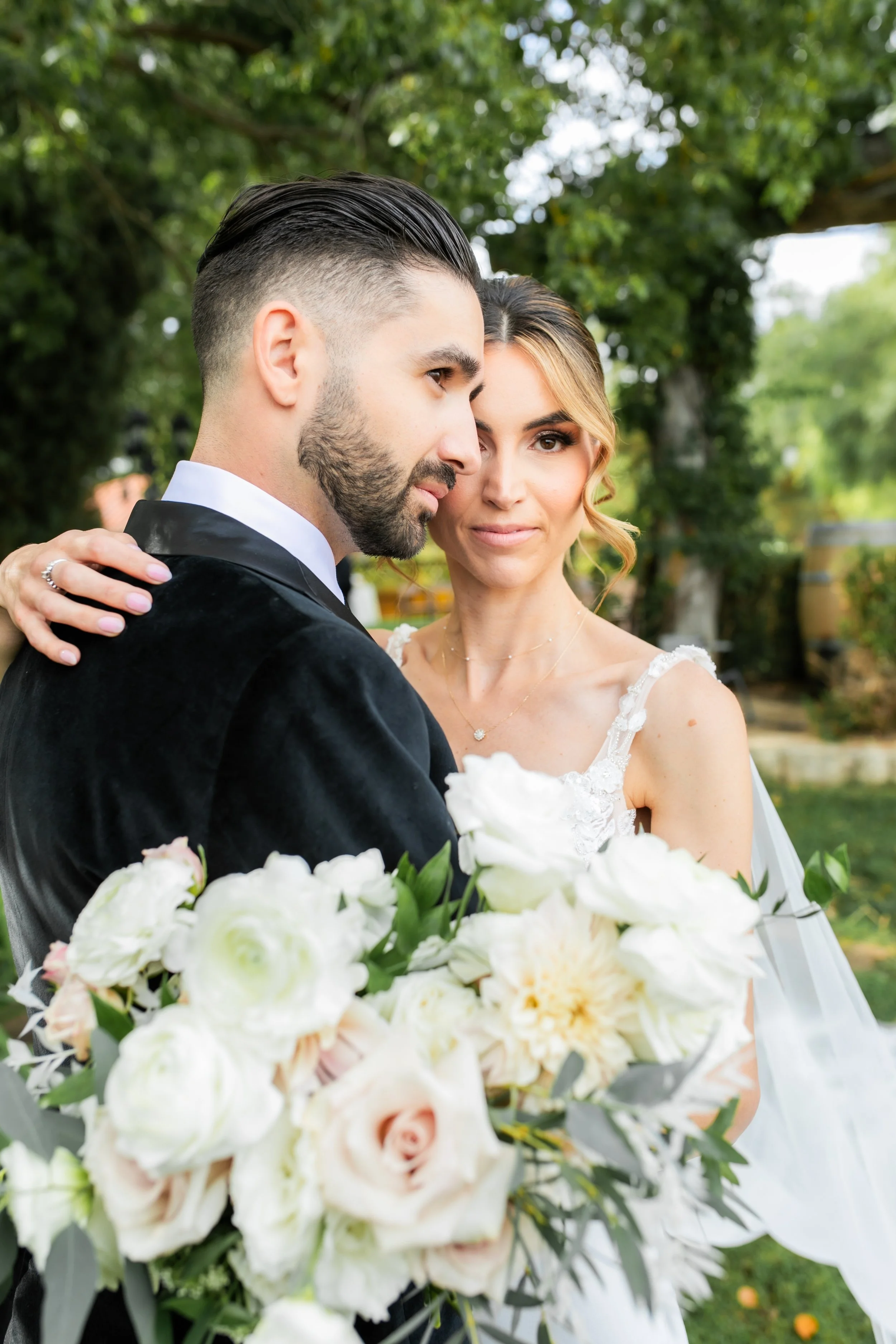 A bride and groom embrace outdoors, the groom in a black tuxedo and the bride in a white wedding dress, holding a bouquet of white and blush flowers, with green trees in the background.