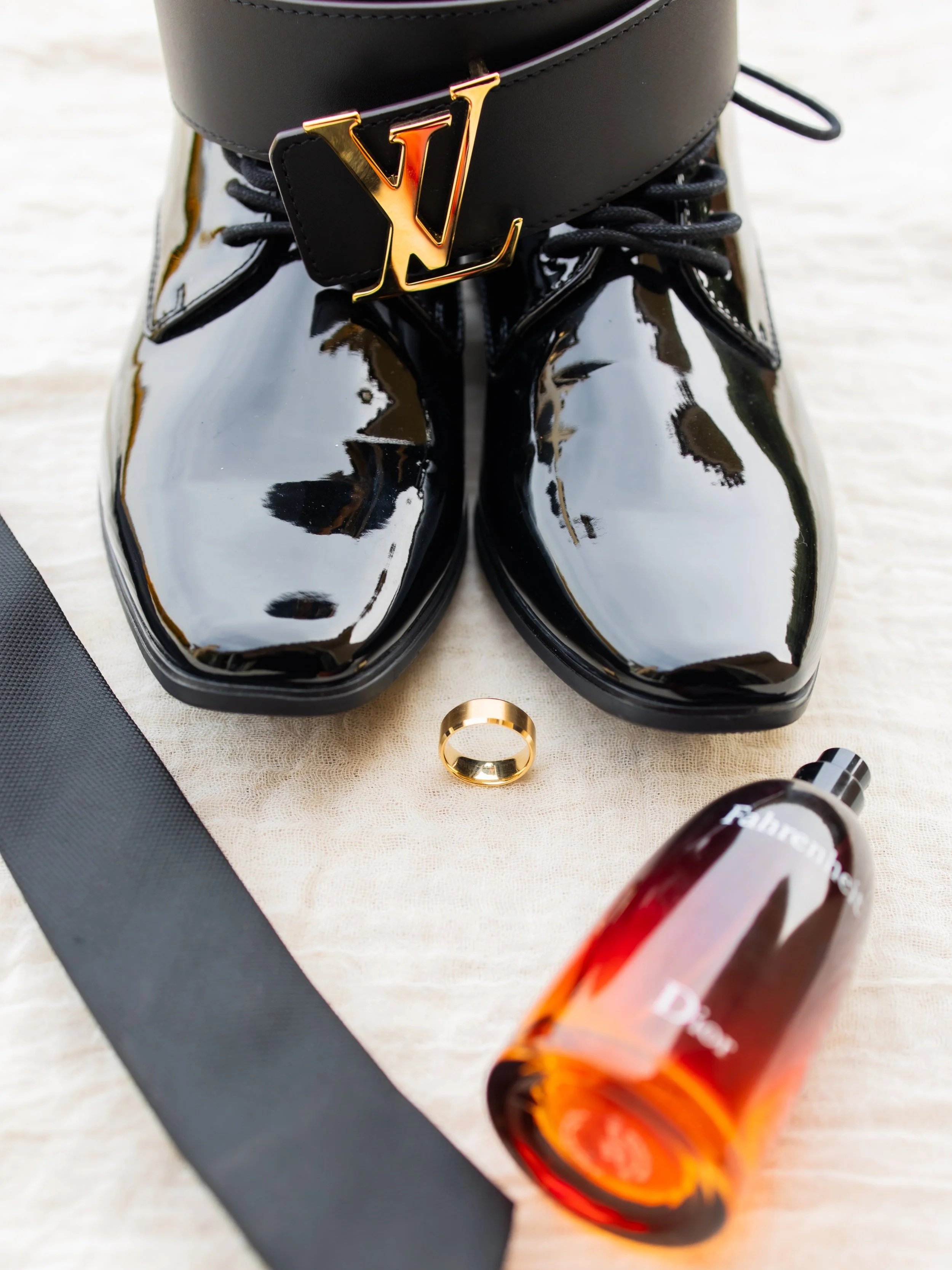 Black shiny leather shoes with black laces, a black belt with a gold Louis Vuitton logo buckle, a gold ring, and a bottle of perfume on a light surface.