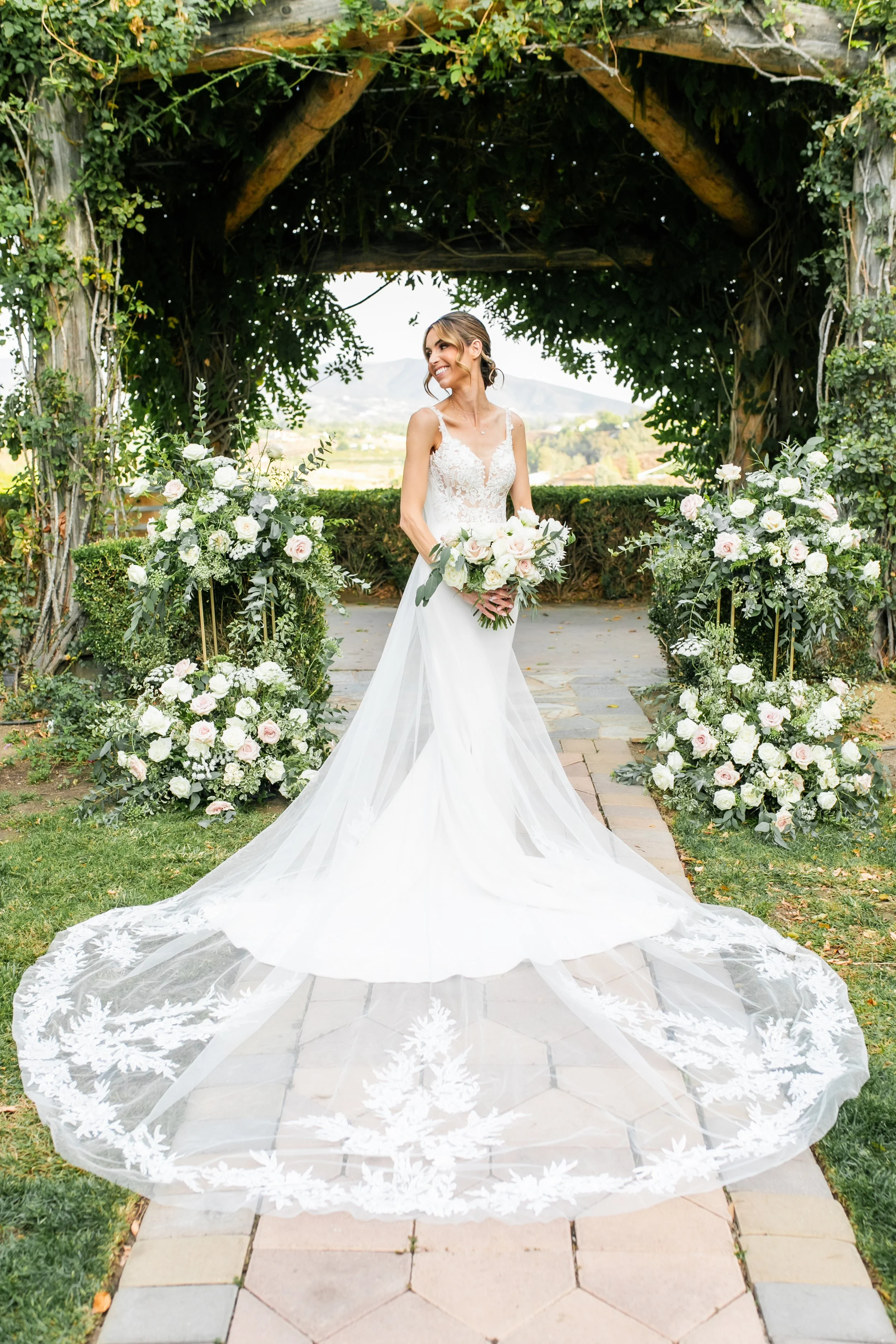 A smiling bride at South Coast Winery in a white wedding gown holding a bouquet of white and pink roses stands under an arch decorated with white flowers and greenery, outdoors on a stone pathway with a garden and landscape in the background.