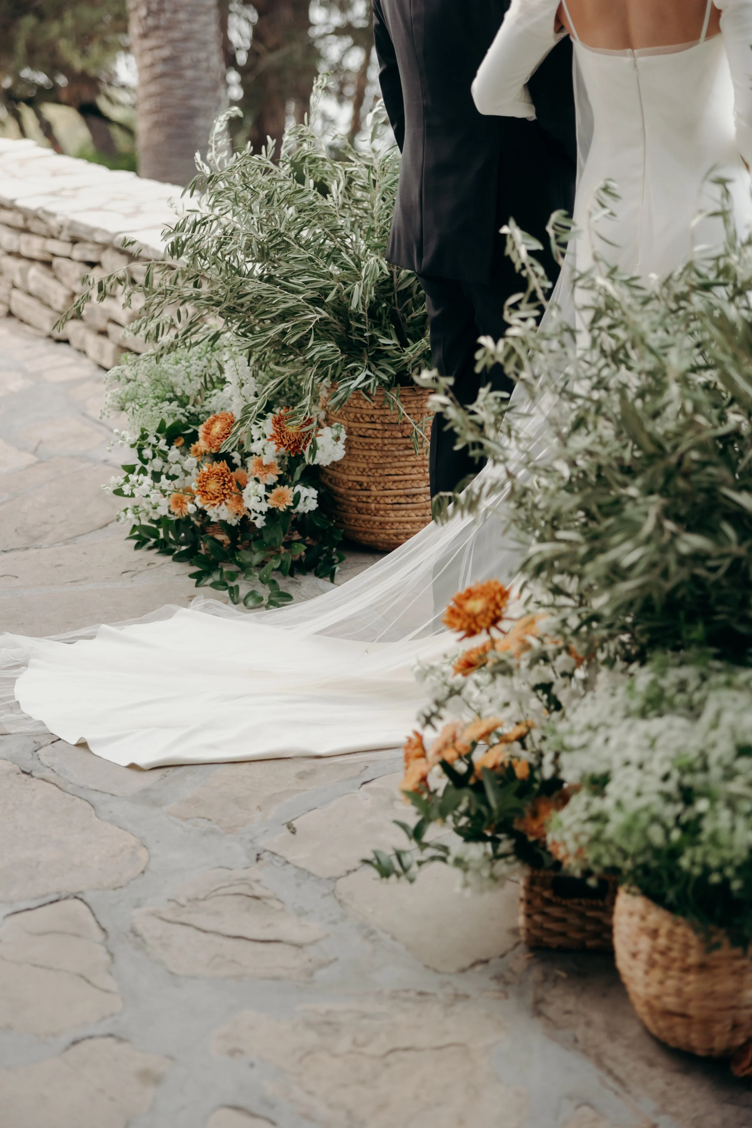 Close-up of a wedding scene showing the bride and groom standing side by side on a stone pathway, surrounded by potted and floral arrangements with greenery and orange and white flowers.