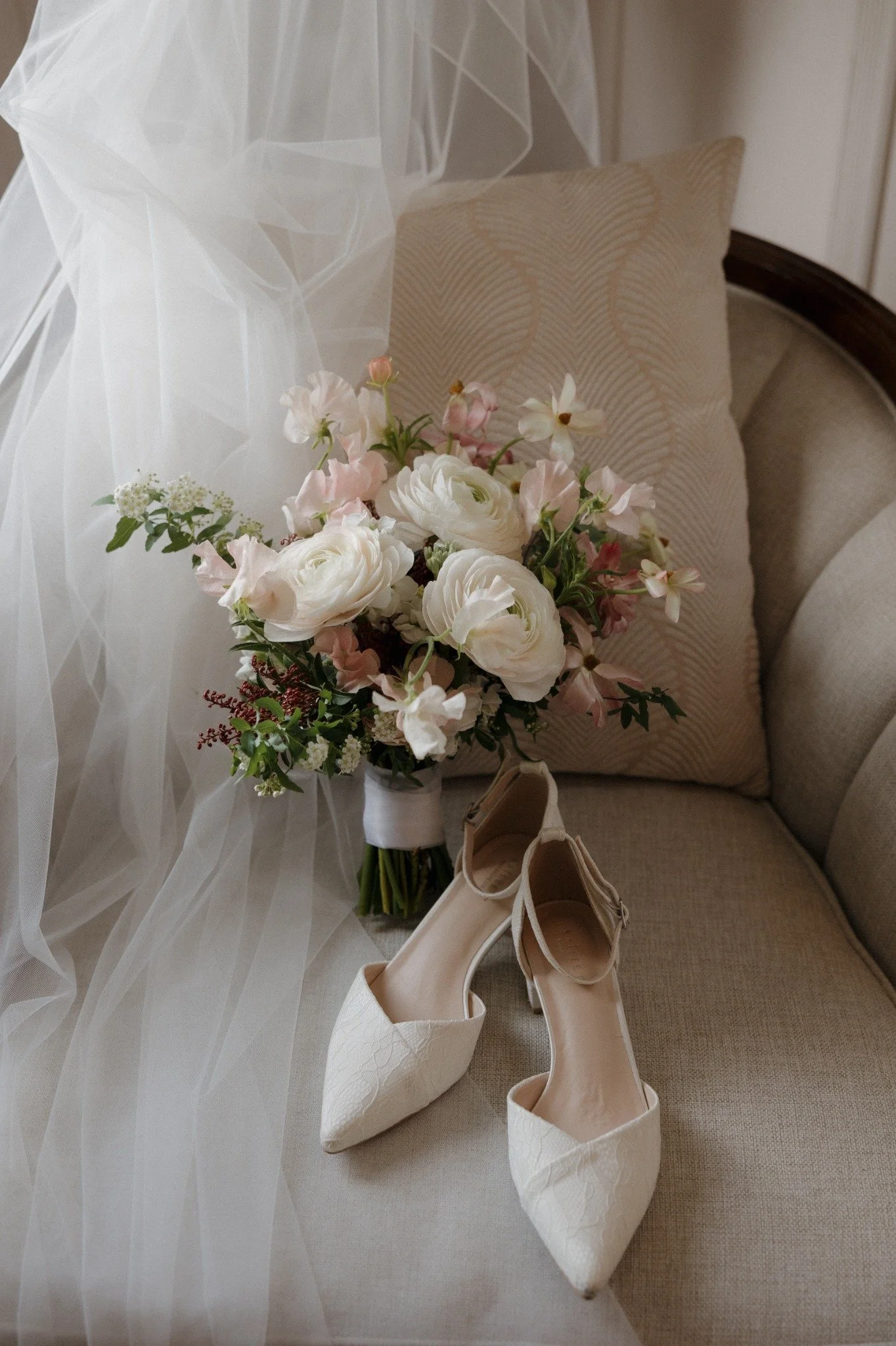 A bouquet of pink and white flowers, a pair of ivory high-heeled shoes, a cushion, and sheer fabric on a beige sofa.