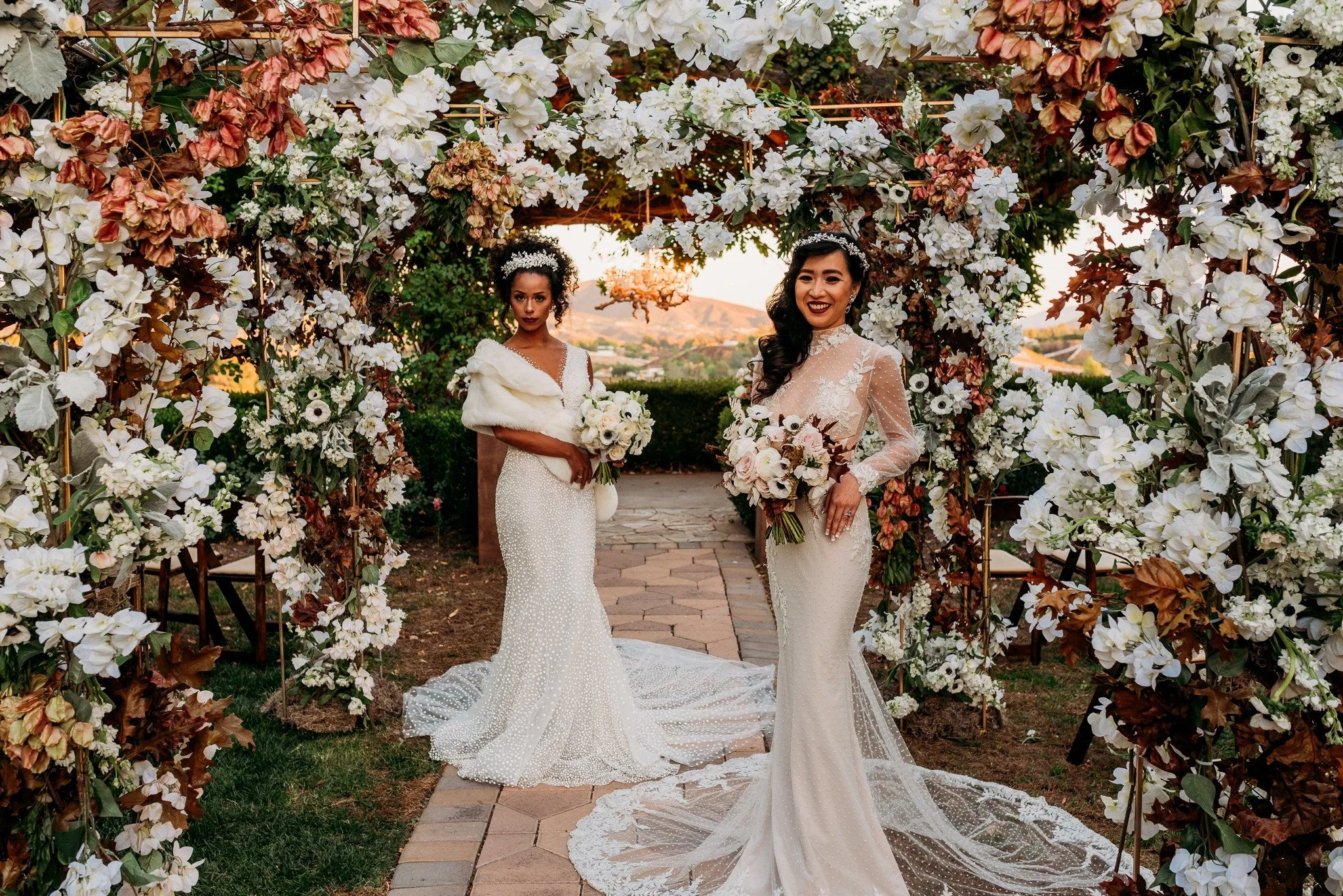 Two brides standing beneath floral ceremony arch at South Coast Winery fall vineyard wedding editorial
