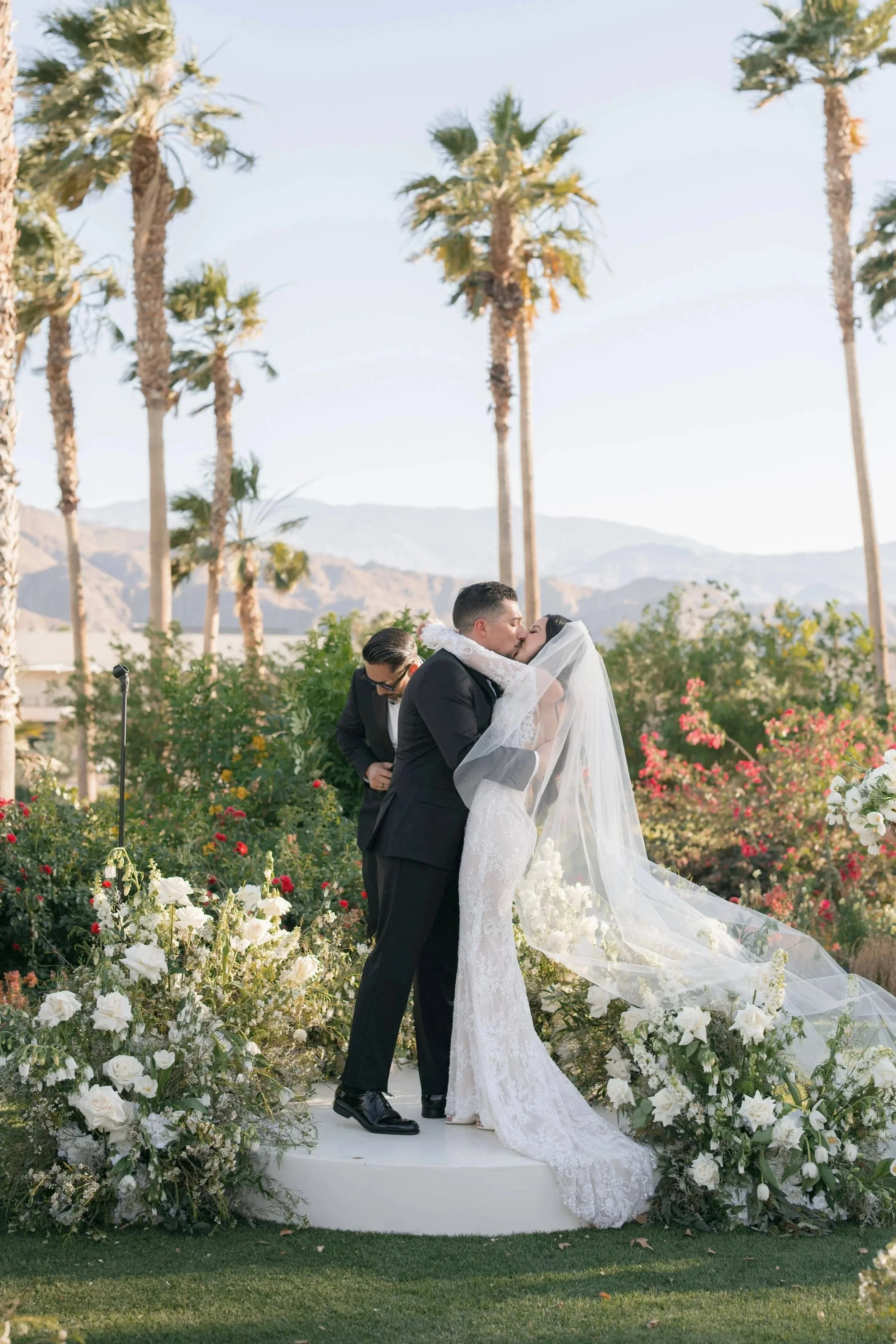Couple sharing a kiss during a wedding ceremony outdoors, with a bridesmaid or groomsman standing nearby, surrounded by floral arrangements and palm trees.