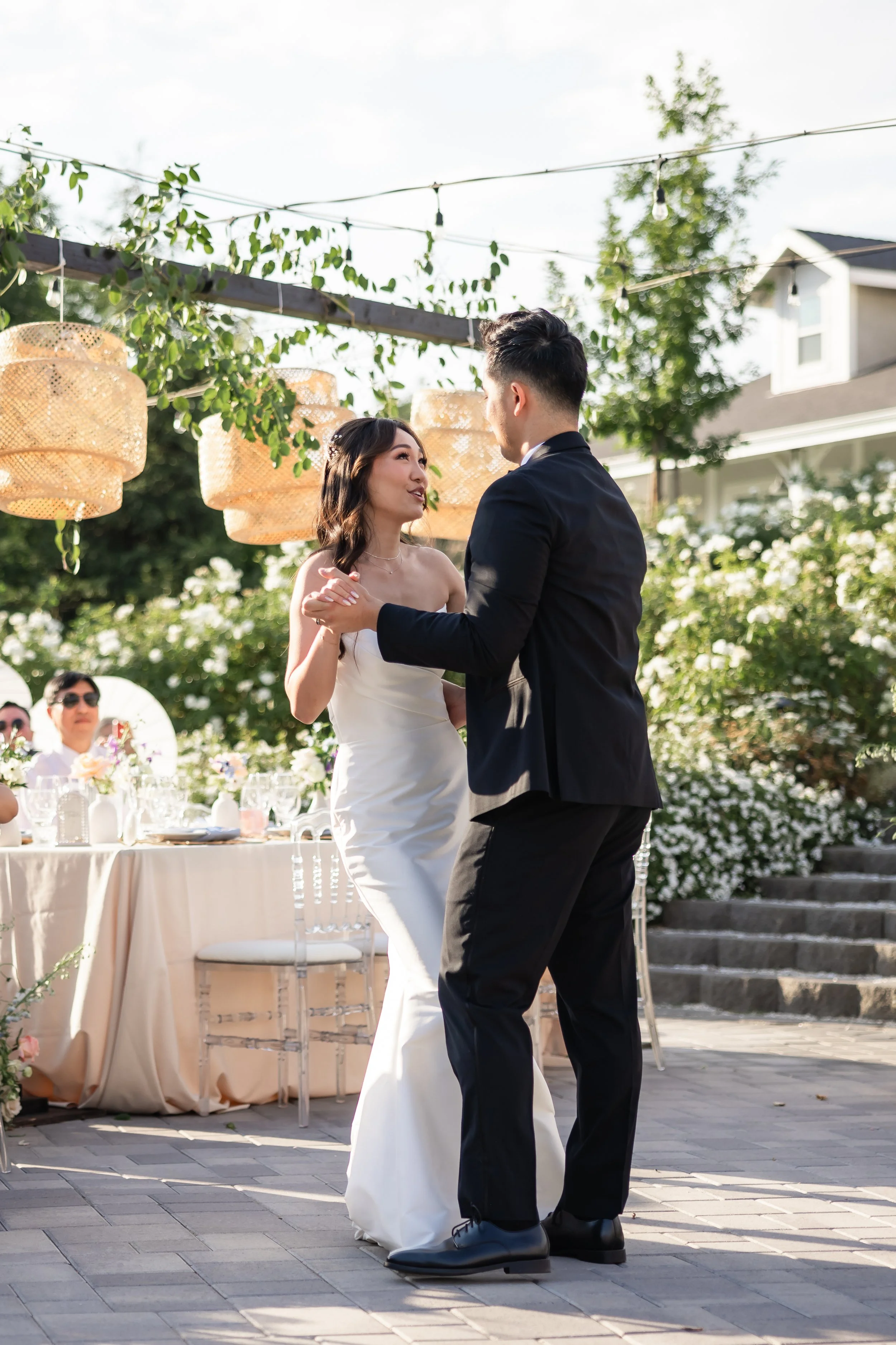 A bride and groom dancing at their outdoor wedding reception during daytime, with guests and decorated tables in the background.