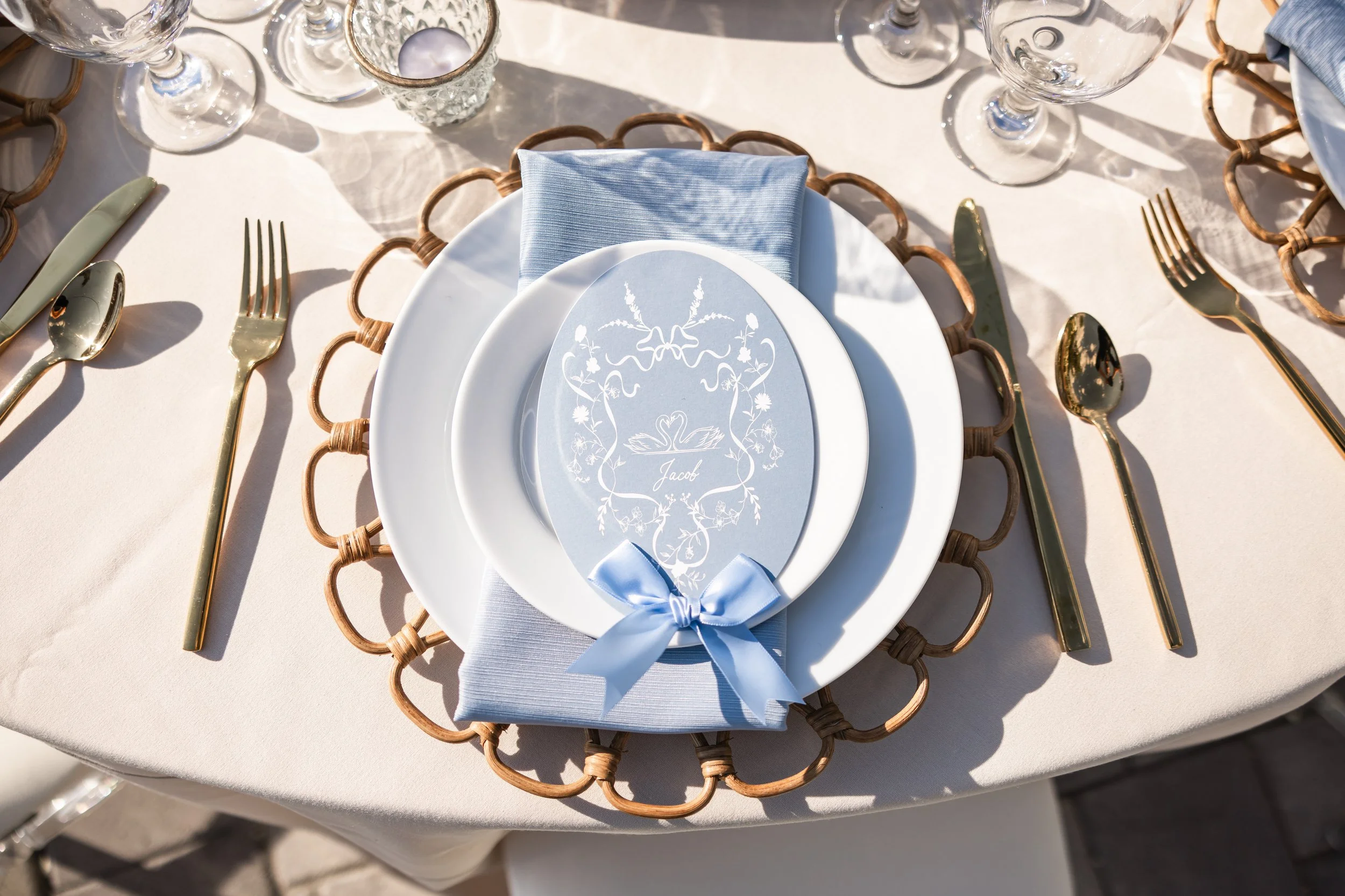 Elegant place setting at a wedding reception with gold utensils, a blue napkin, and a blue printed menu on top of white plates, all arranged on a white tablecloth.