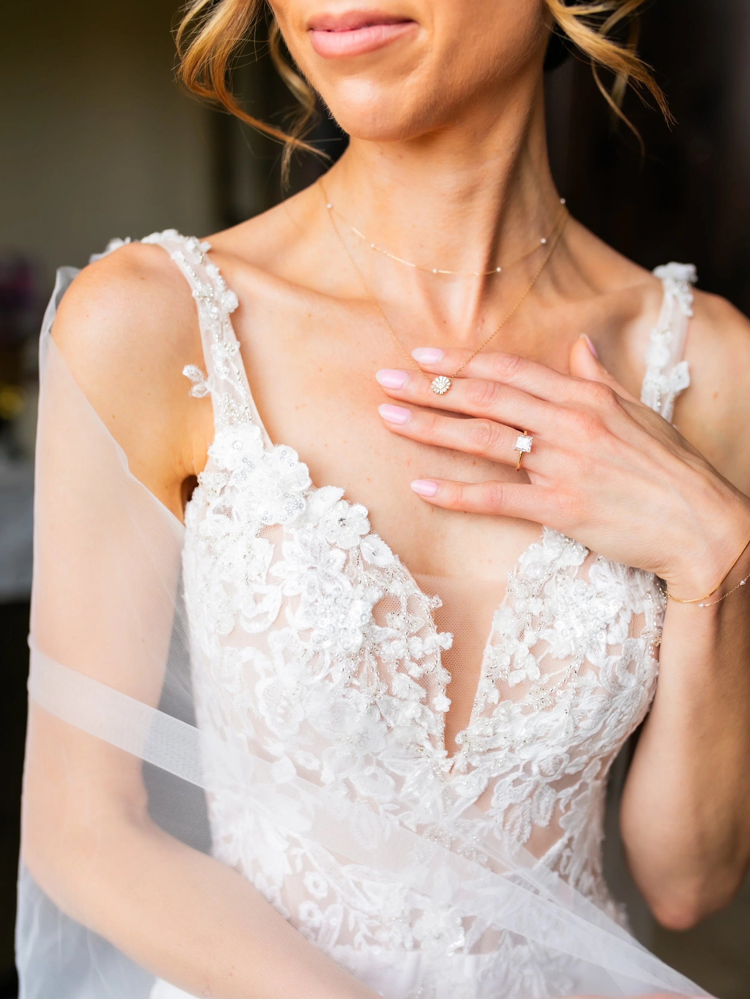Close-up of a woman wearing a white lace wedding dress, displaying jewelry including a necklace and rings, with her hand on her chest.