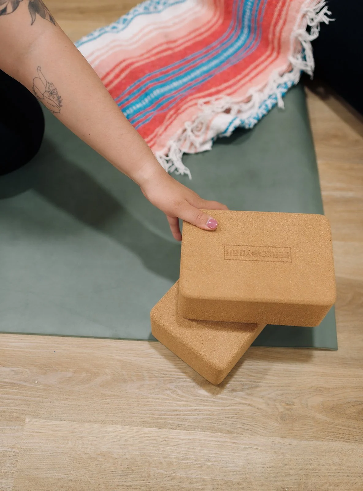 Person with a tattoo on their arm pressing two cork yoga blocks on a wooden floor with a futon or mat with a multicolored blanket on top.
