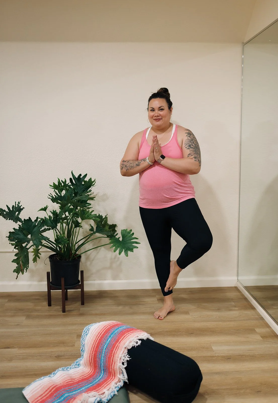 A woman standing in a yoga pose inside a room, wearing a pink tank top and black leggings, with tattoos on her arms, a watch, and an Apple Watch, next to a large green plant and a rolled-up towel on the floor.