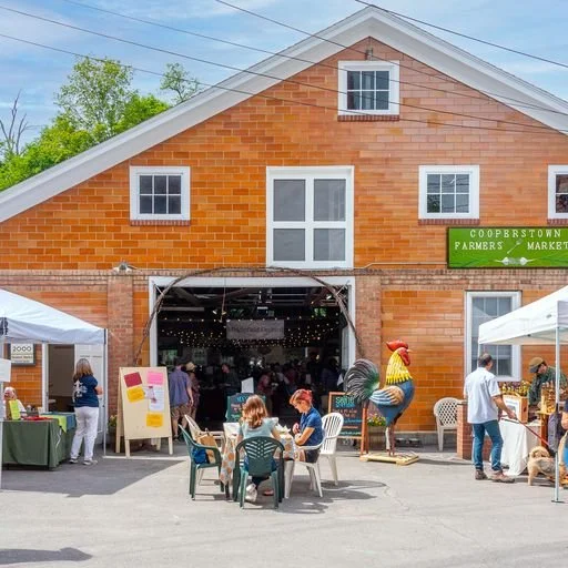 Outside of Farmers' Market, people sitting at tables, looking at booths, view to the indoor Market